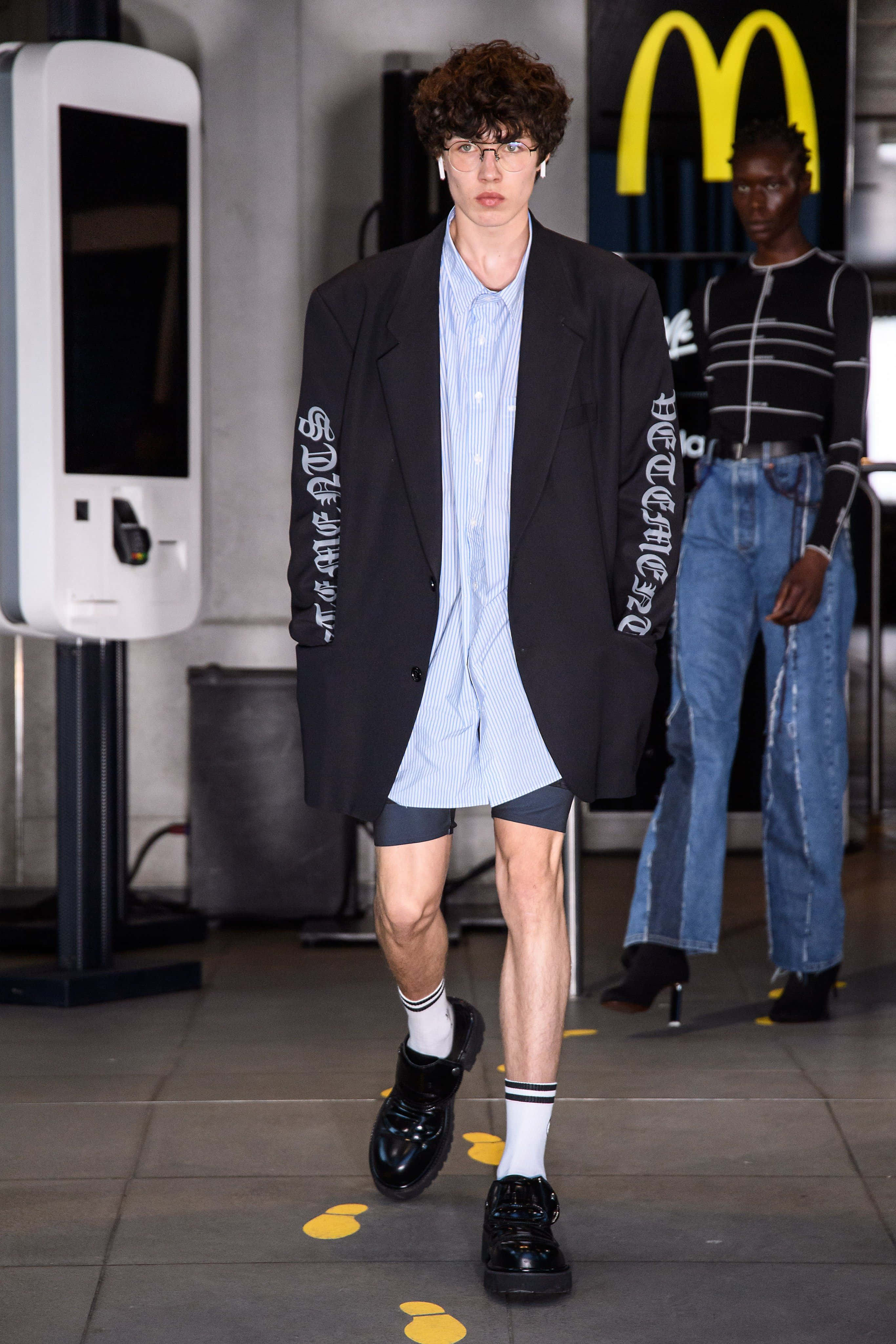 穿上 Vetements，感受活力與生動 壁紙 — 一位模特兒自信地穿著帶有白色字樣的黑色西裝外套，內搭條紋襯衫和短褲，在背景有麥當勞標誌的T台上行走。