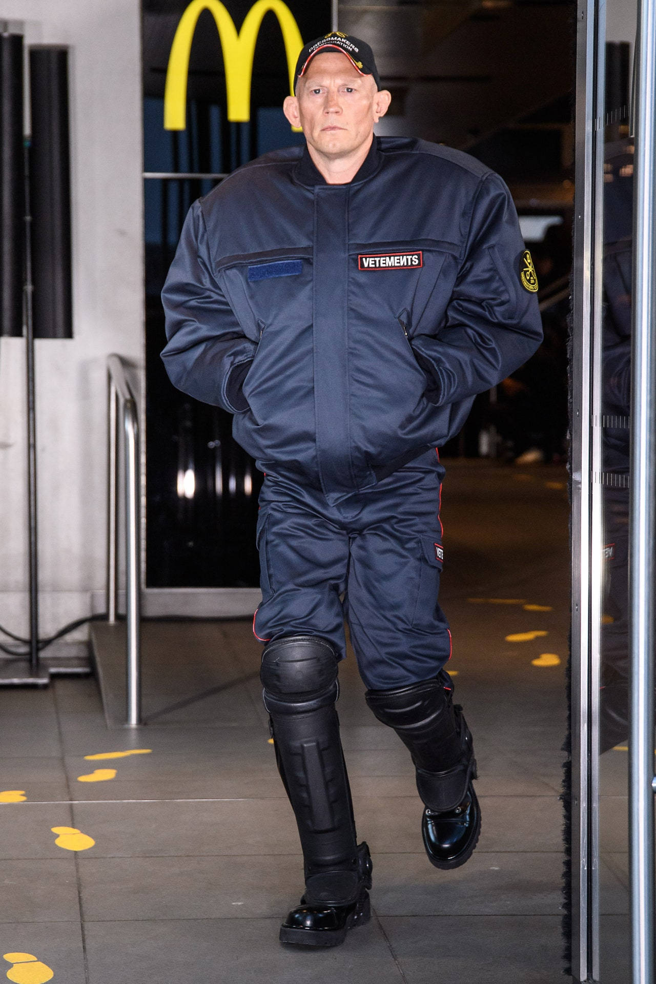 Vetements 时装周在麥當勞舉行 壁紙 — 一名身穿深藍色VETEMENTS連身衣和黑色護膝的男子走過麥當勞入口，地板上有黃色腳印。