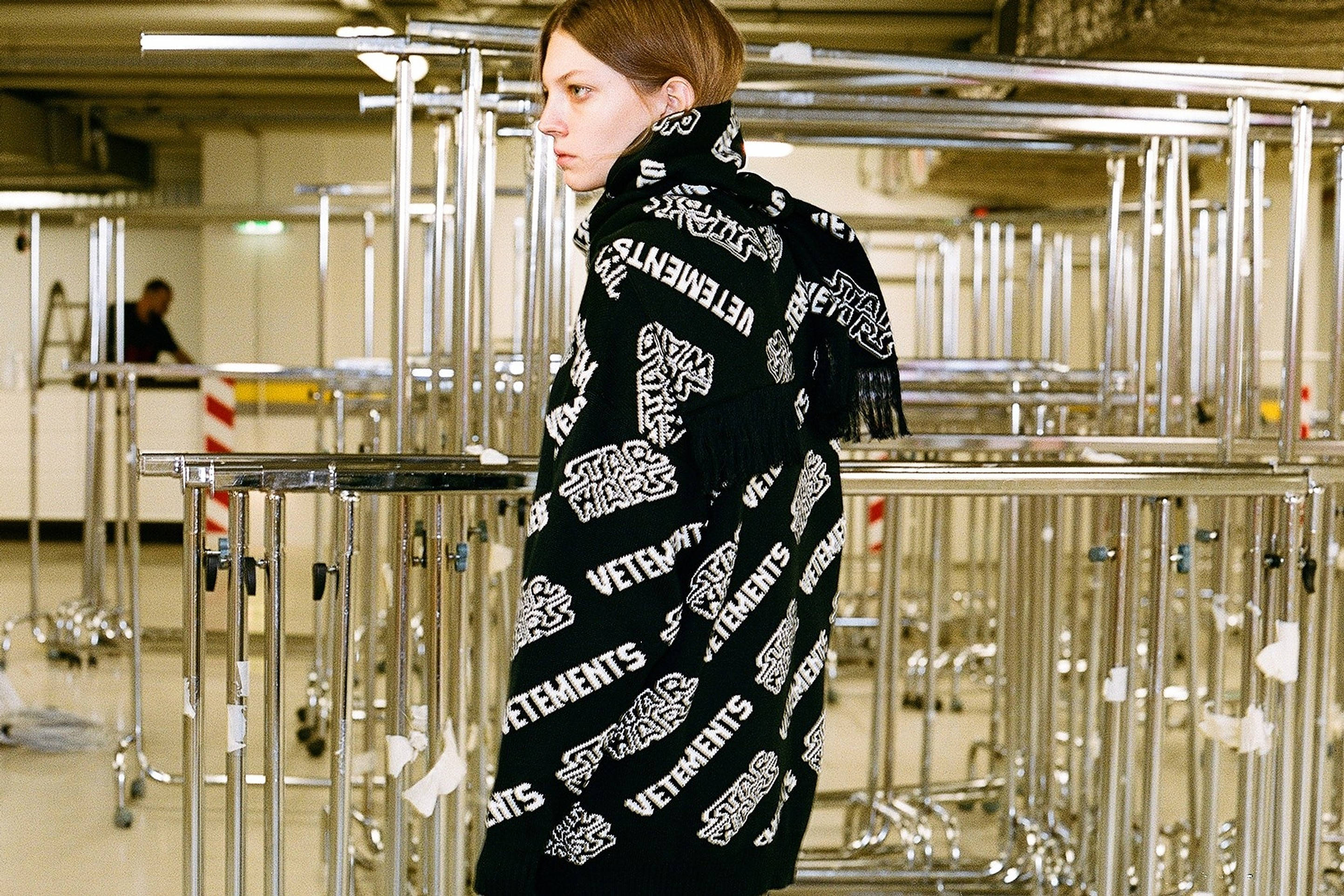 Vetements x 星際大戰 壁紙 — 一個人披著黑色Vetements圍巾，上面有白色重複標誌，站在倉庫式空間的鉻製欄杆之間。