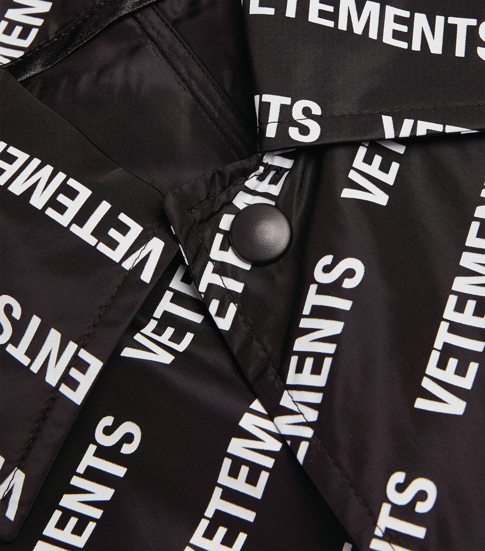 Vetements 服裝品牌 壁紙 — 黑色外套的特寫，布料上對角重複印有粗體白色「VETEMENTS」文字，並有一顆黑色鈕扣。