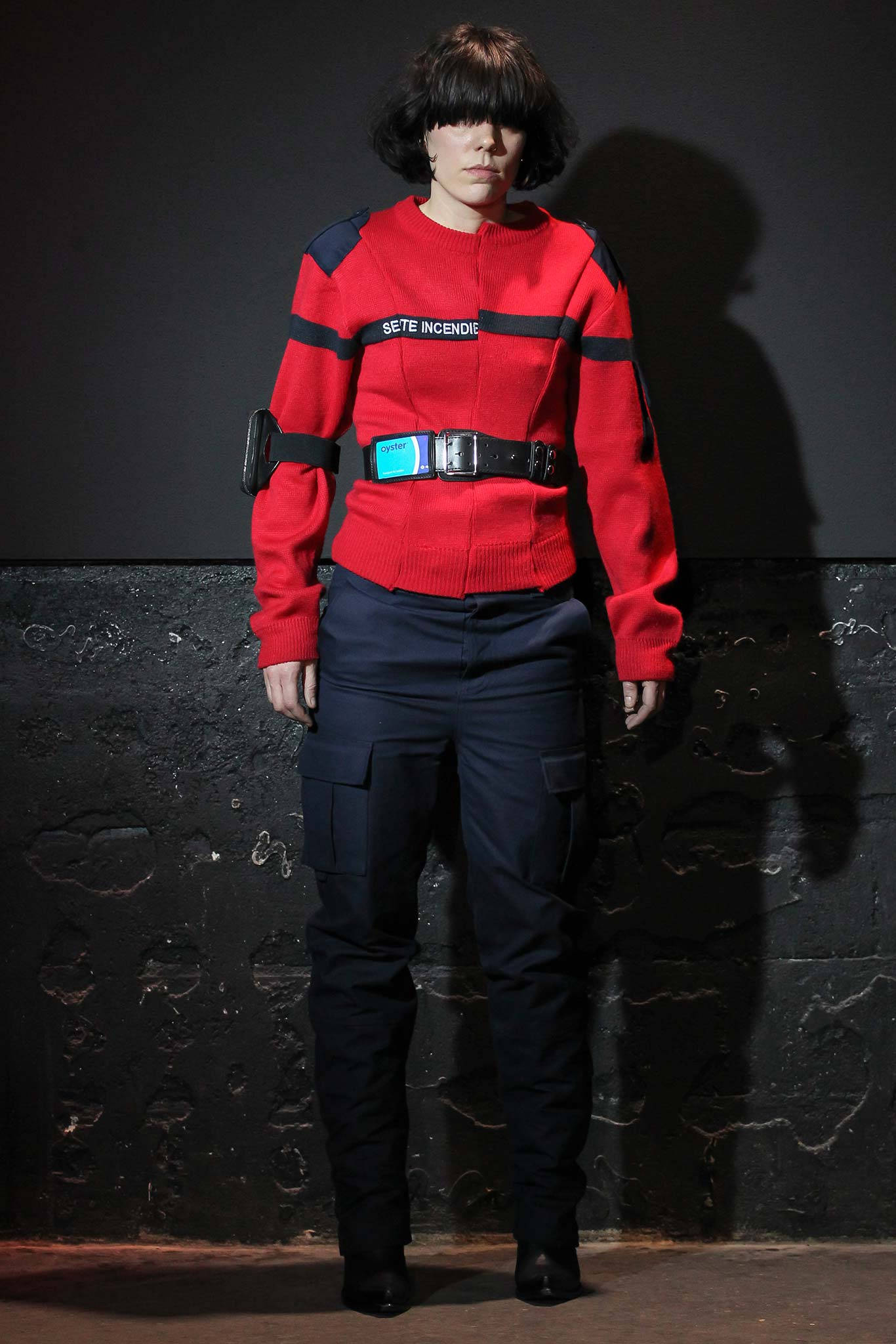 Vetements 紅色長袖上衣 壁紙 — 一個人穿著大膽的紅色毛衣，帶有黑色條紋和文字，站在深色紋理牆前，投射出強烈的陰影。