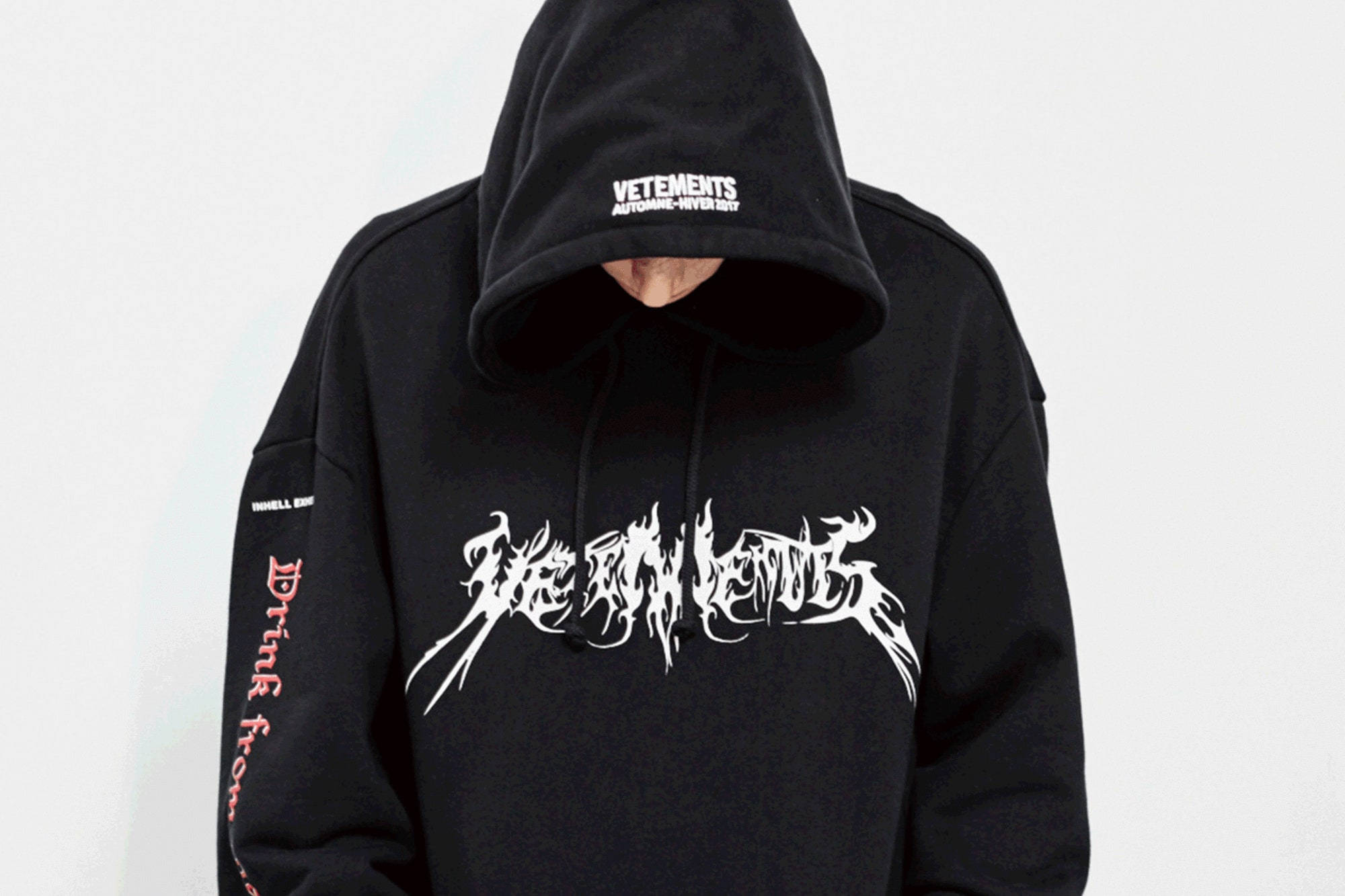 Vetements 全黑連帽衫 壁紙 — 一個人穿著黑色連帽衫，上面有白色塗鴉文字和紅色袖子字母，臉藏在帽子下，背景是純白色。