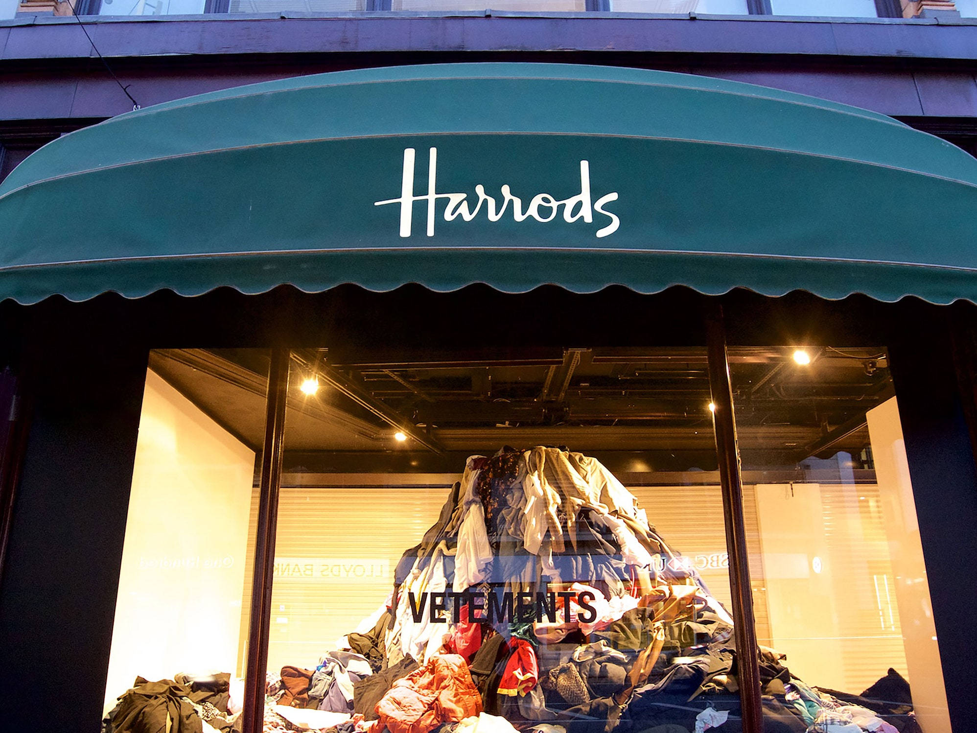 Vetements 於哈羅德百貨 壁紙 — 在昏暗的商店櫥窗中，一堆色彩繽紛的衣物在綠色Harrods遮陽篷下形成如山般的展示。
