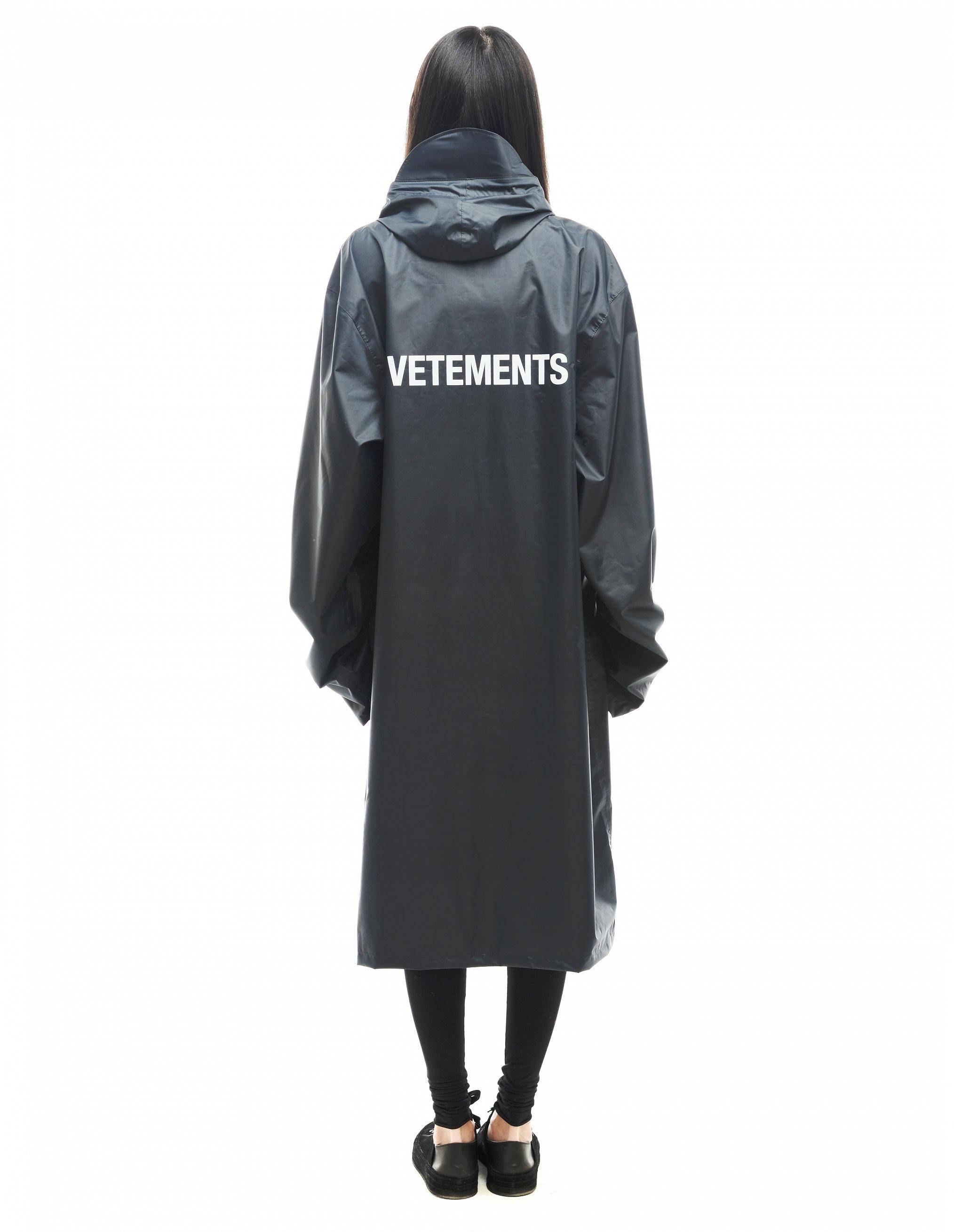 Vetements 雨衣 壁紙 — 從背後看到的一個人穿著一件長黑色連帽雨衣，背面有醒目的白色VETEMENTS品牌標誌。