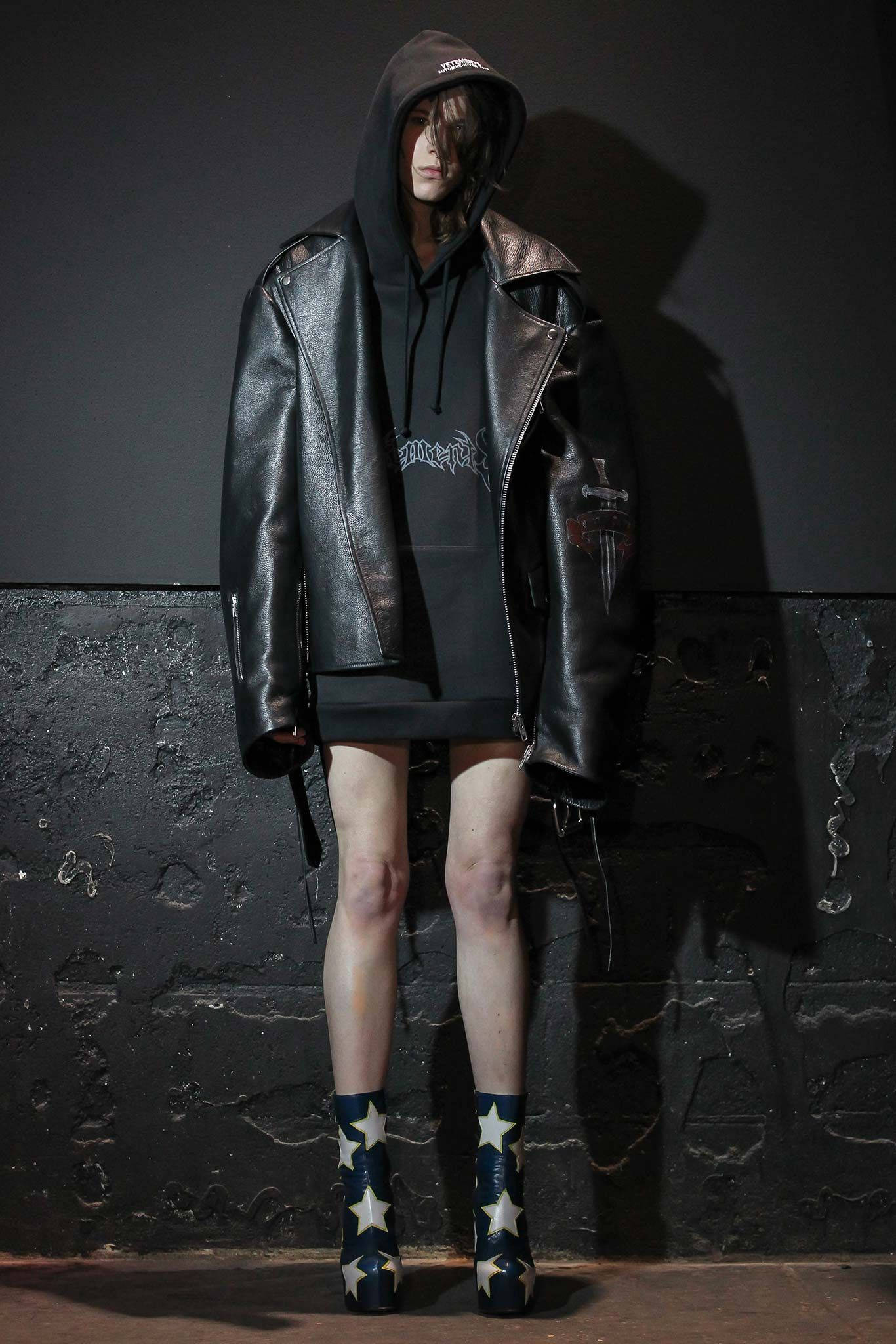 Vetements 黑色皮革大衣 壁紙 — 一個人穿著黑色皮褸和星形圖案靴子，在戲劇性燈光下靠著深色紋理牆壁站立。
