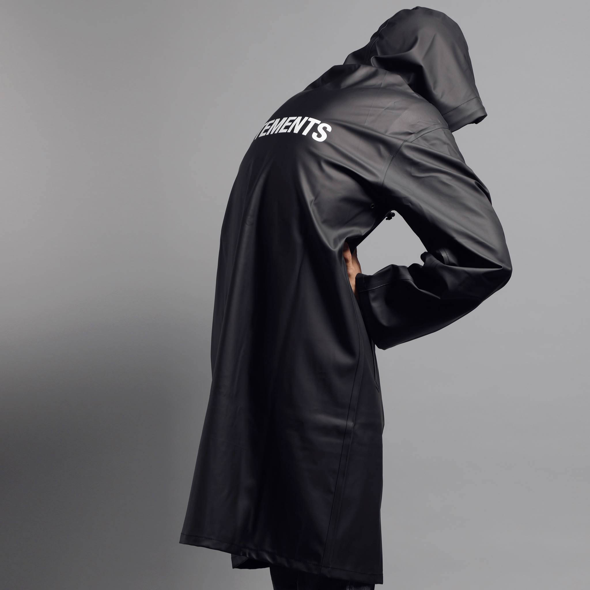 Vetements 黑色雨衣 壁紙 — 一個人穿著黑色連帽雨衣，背面有白色文字，站在純灰色背景前。