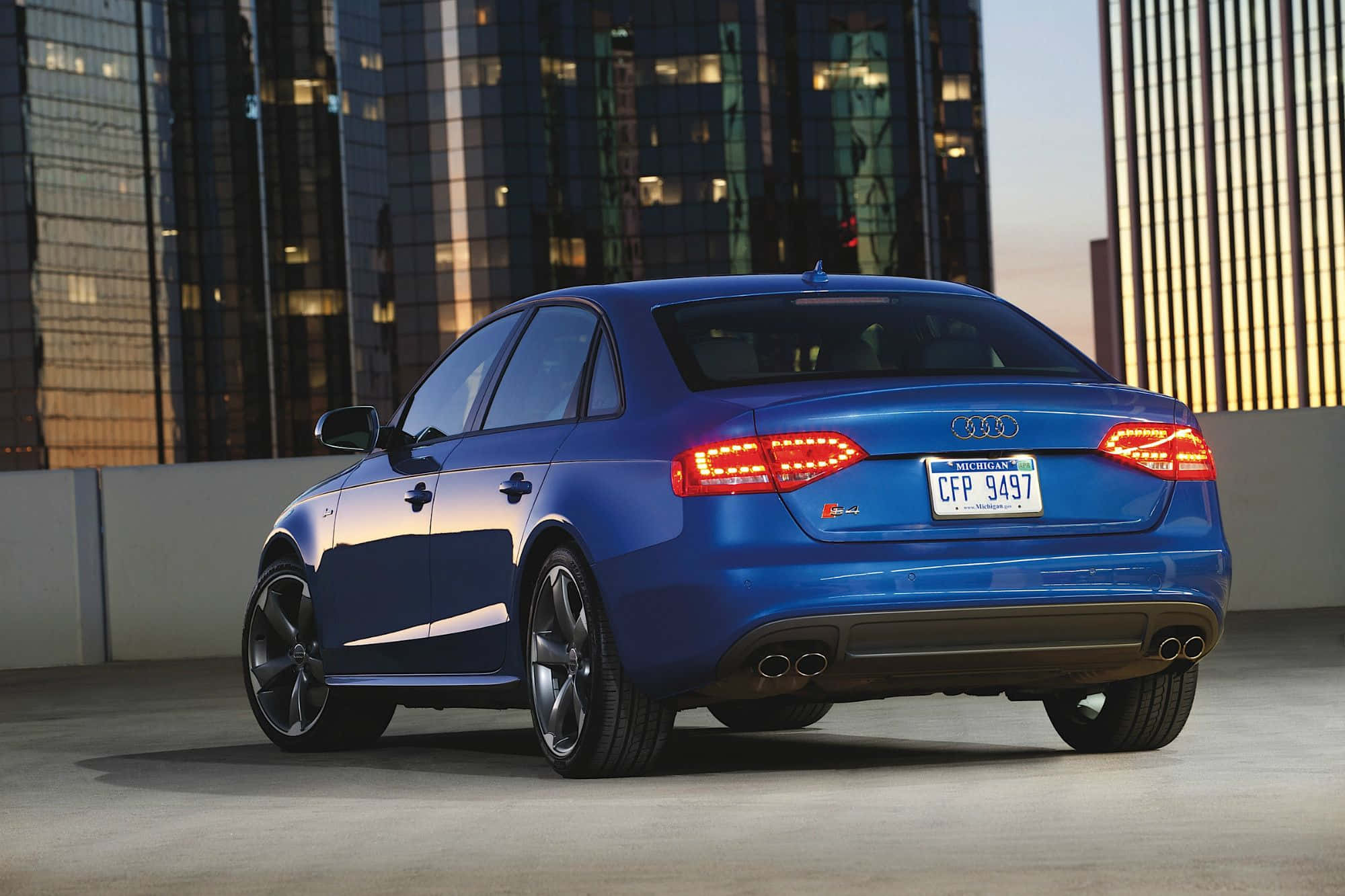 Audi S4 桌布 壁紙 — 一輛充滿活力的藍色奧迪轎車在金色夕陽下閃閃發光，停放在屋頂上，背景是高聳的玻璃摩天大樓。