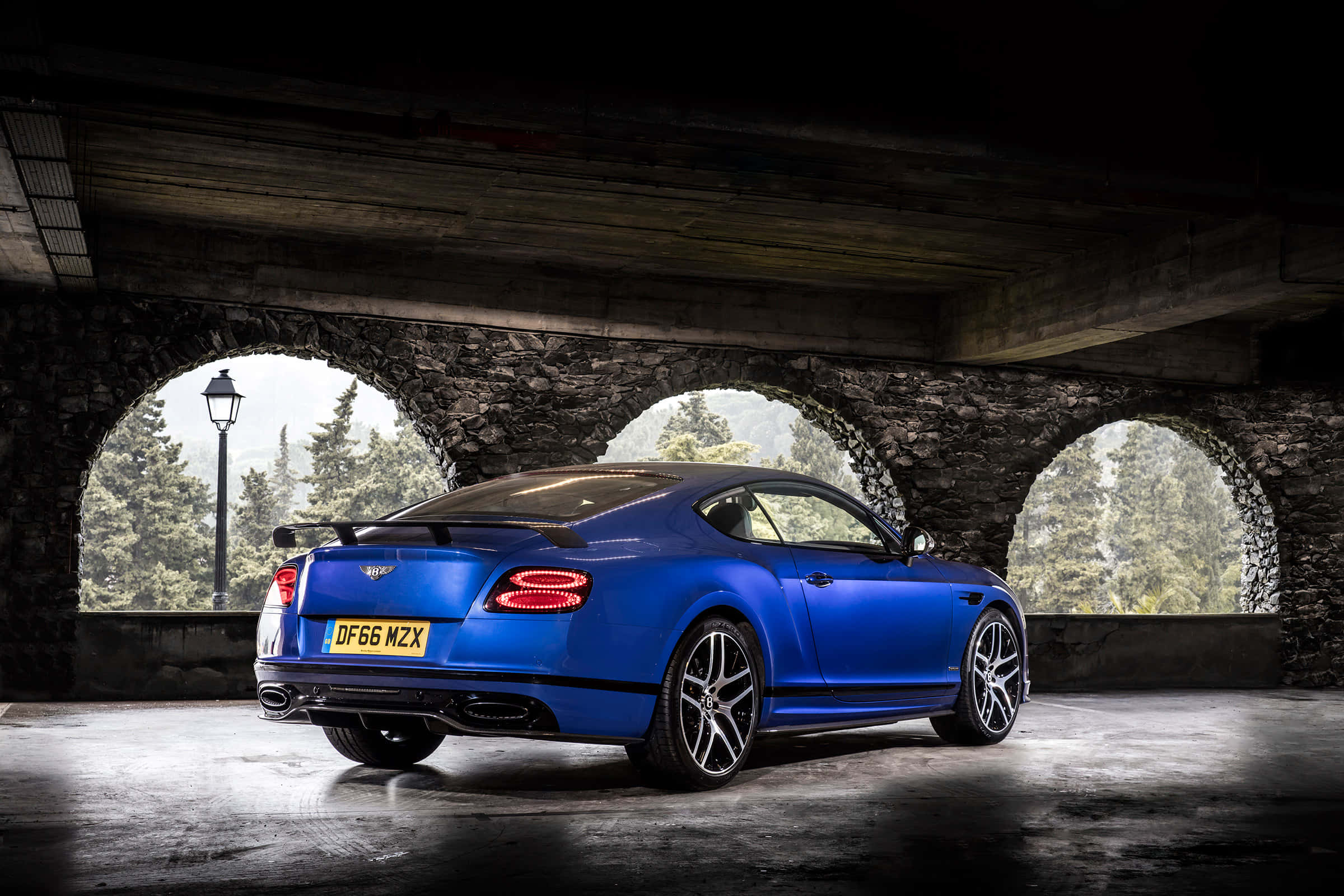 Bentley Continental Supersports 桌布 壁紙 — 一輛充滿活力的藍色賓利 Continental GT 在石拱門下，在戲劇性的燈光下閃耀，並可欣賞森林景色。