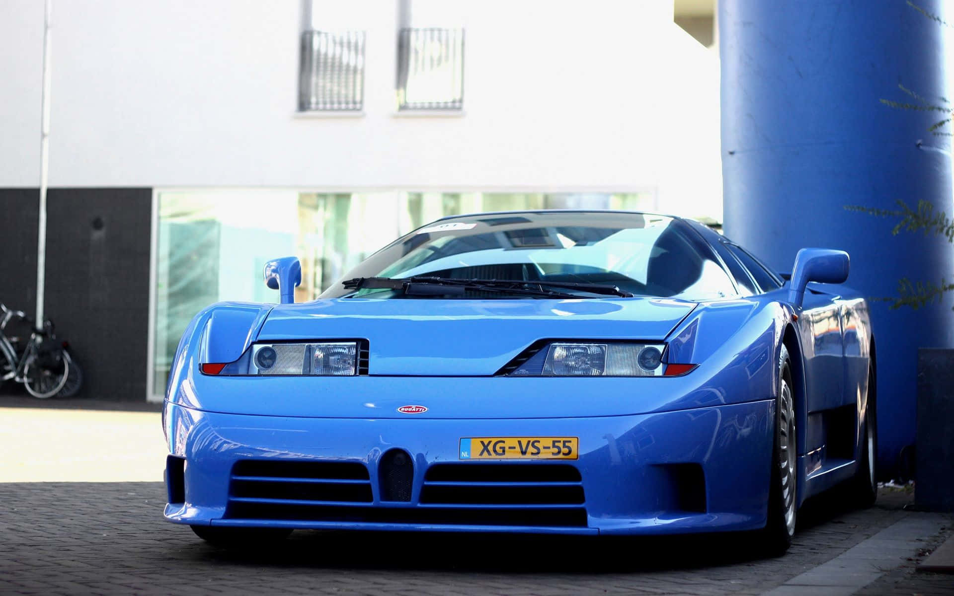 Bugatti Eb110 桌布 壁紙 — 一輛鮮藍色Bugatti EB110超級跑車在日光下閃閃發光，停泊在現代建築旁的藍色柱子旁邊。