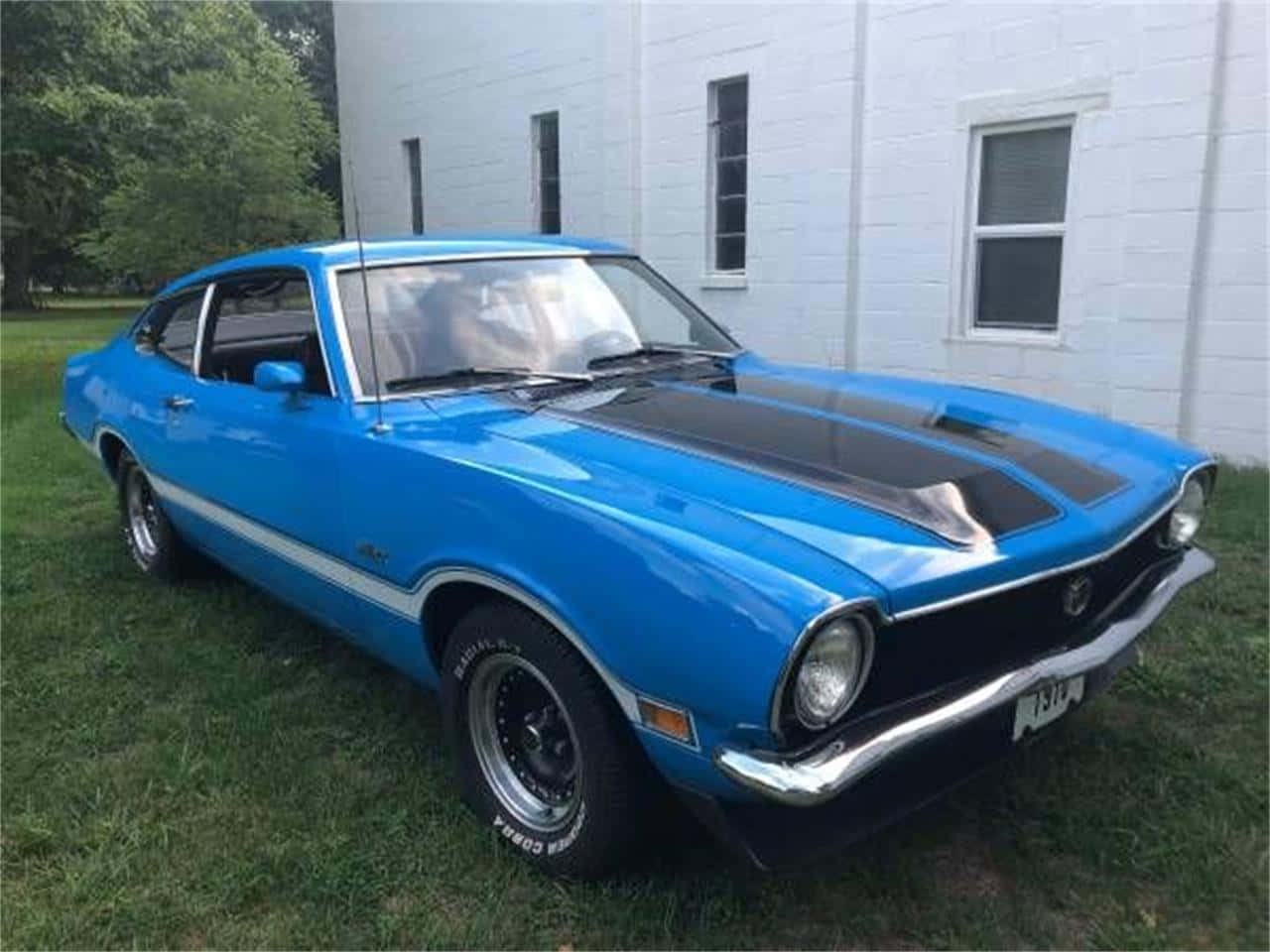 Ford Maverick 桌布 壁紙 — 一輛充滿活力的藍色經典肌肉車，帶有黑色引擎蓋條紋，停在白色磚砌建築旁的草地上。