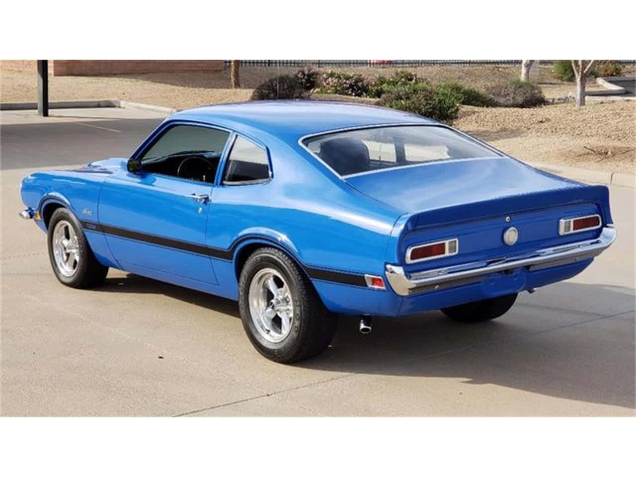 Ford Maverick 桌布 壁紙 — 一輛充滿活力的藍色經典肌肉車，帶有黑色賽車條紋，停在陽光明媚的混凝土停車場上。