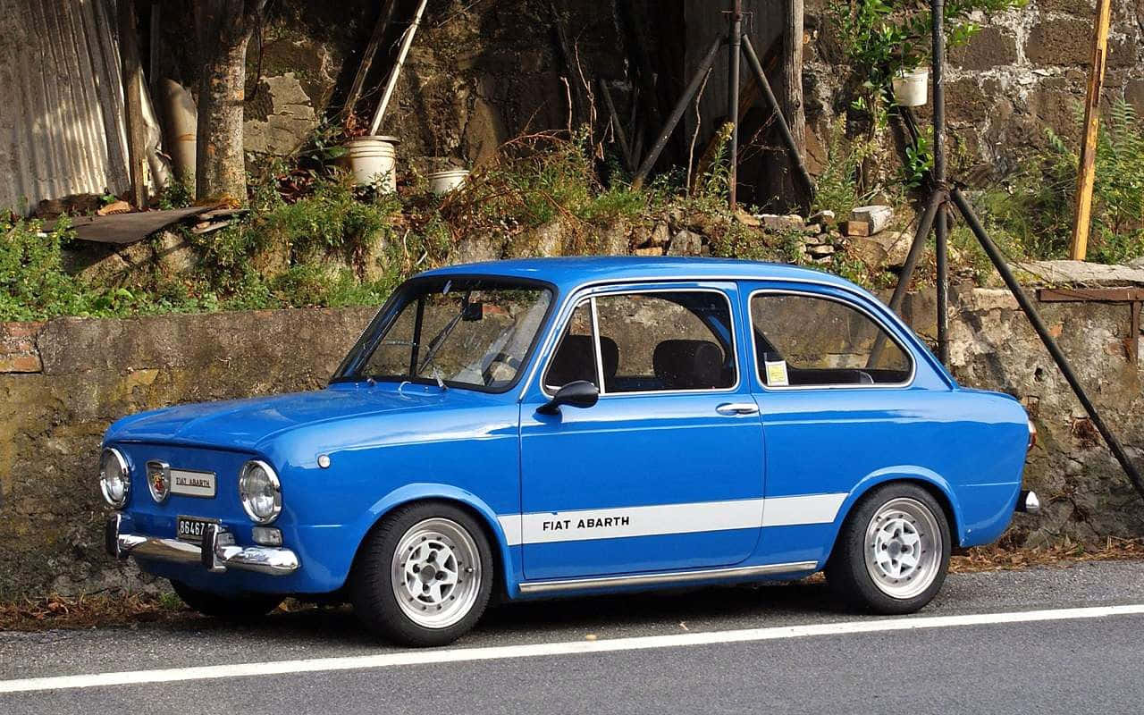 Fiat 500l 桌布 壁紙 — 一輛充滿活力的藍色 Fiat Abarth 經典汽車停泊在路邊，旁邊是帶有綠色植物的風化石牆。