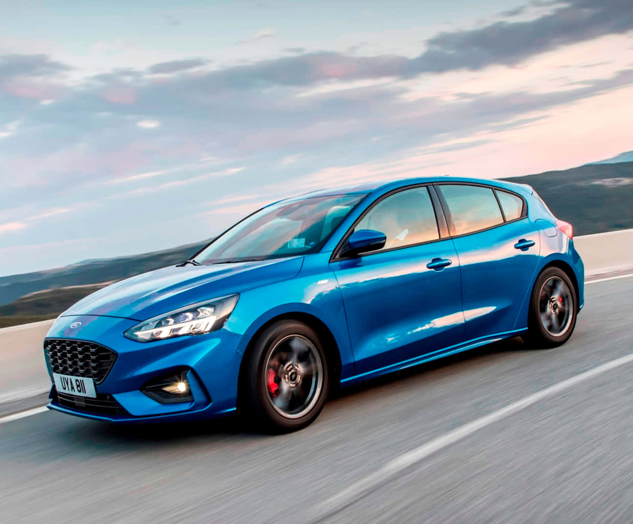 Ford Focus 桌布 壁紙 — 一輛充滿活力的藍色福特福克斯掀背車在柔和粉紅和藍色日落天空下沿著海岸高速公路飛馳。