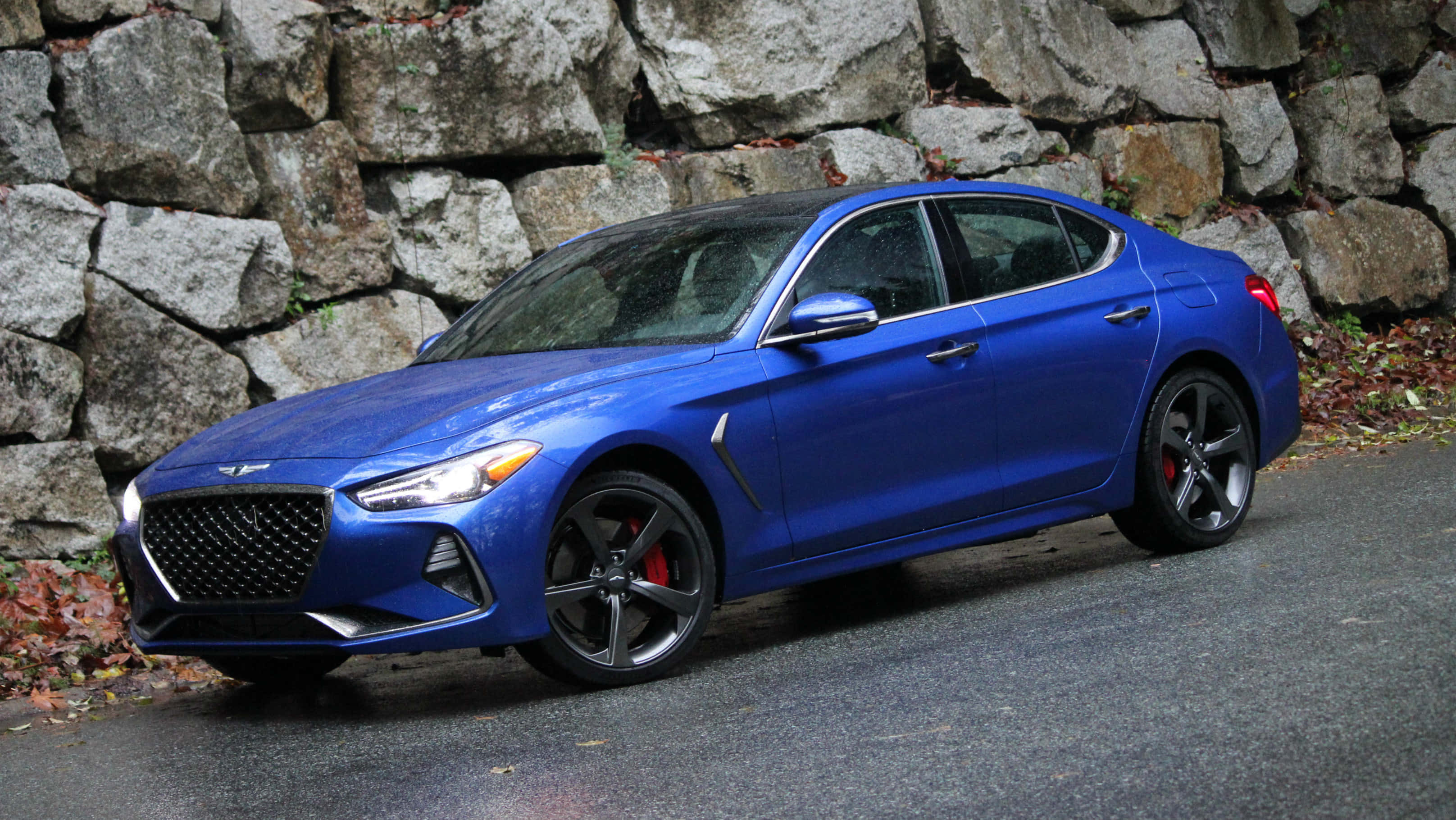 Genesis G70 桌布 壁紙 — 一輛鮮藍色現代Genesis轎車，配備黑色輪圈和紅色制動卡鉗，停在潮濕的道路上，旁邊是長滿苔蘚的石牆。