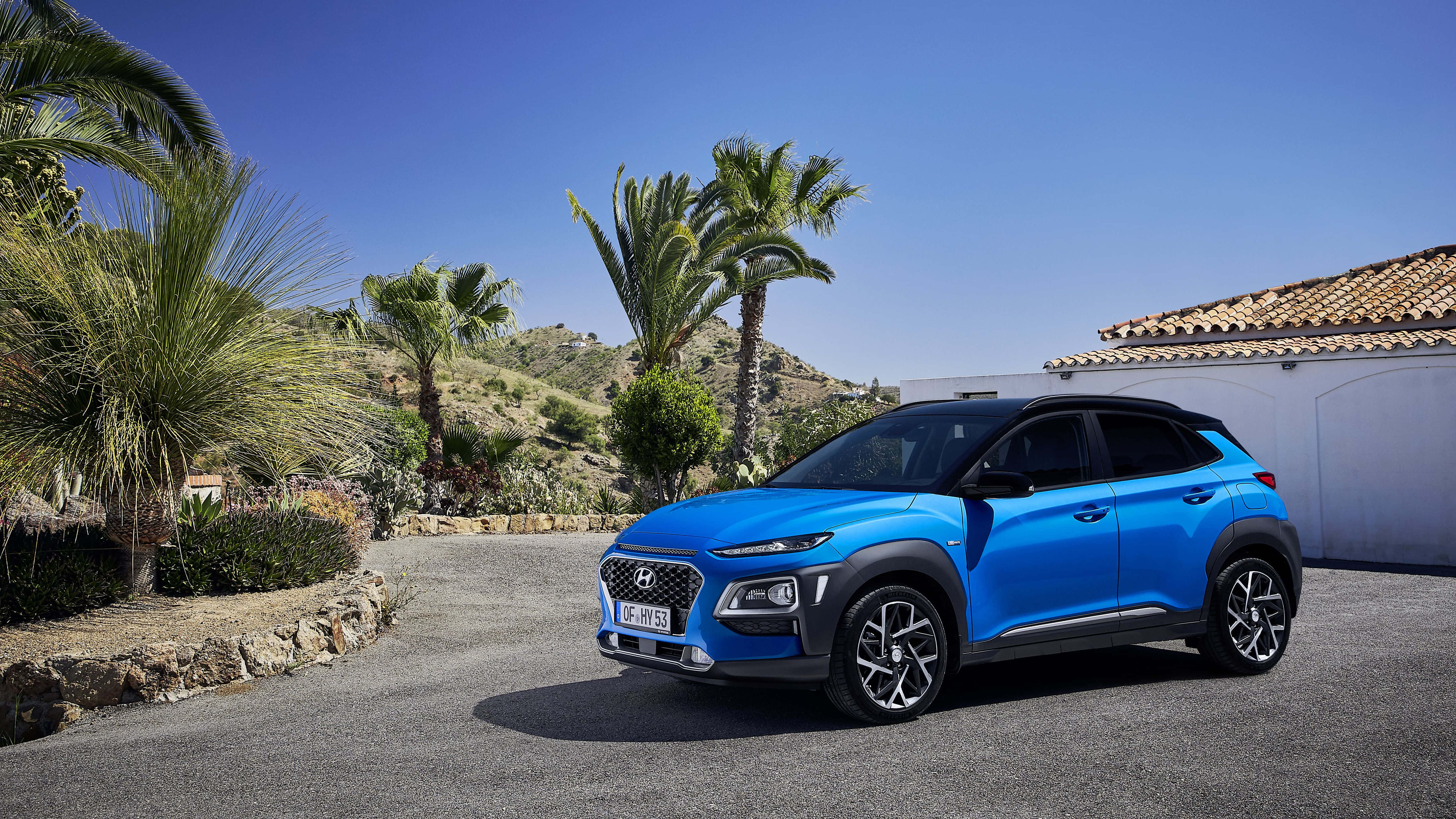 Hyundai Kona 桌布 壁紙 — 一輛充滿活力的藍色現代Kona SUV停泊在陽光普照的車道上，旁邊有棕櫚樹和白色建築，背景是清澈的藍天。