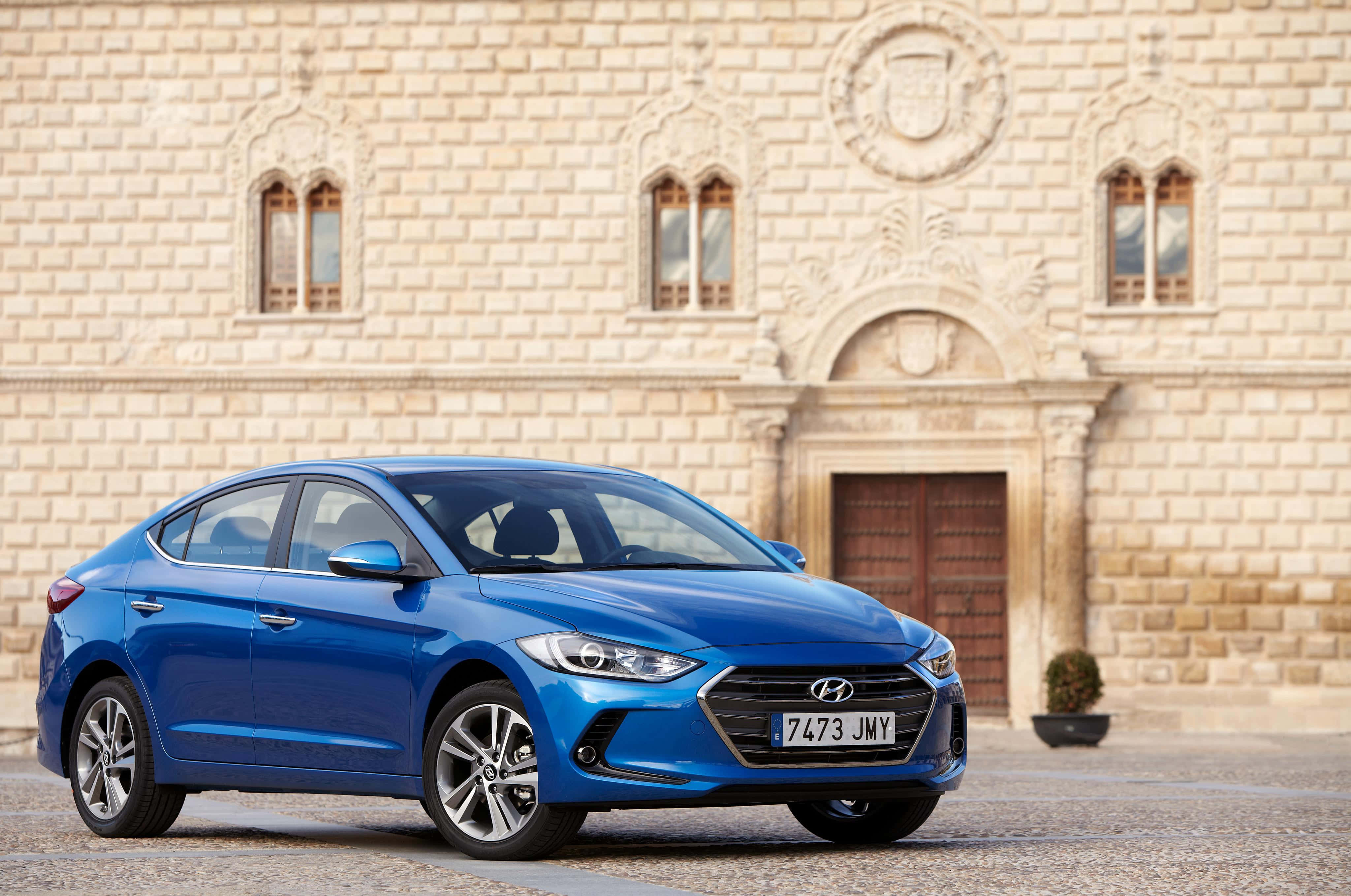 Hyundai Elantra 桌布 壁紙 — 一輛充滿活力的藍色現代轎車在日光下閃閃發光，停在一個帶有拱形窗戶和木門的歷史石建築前。