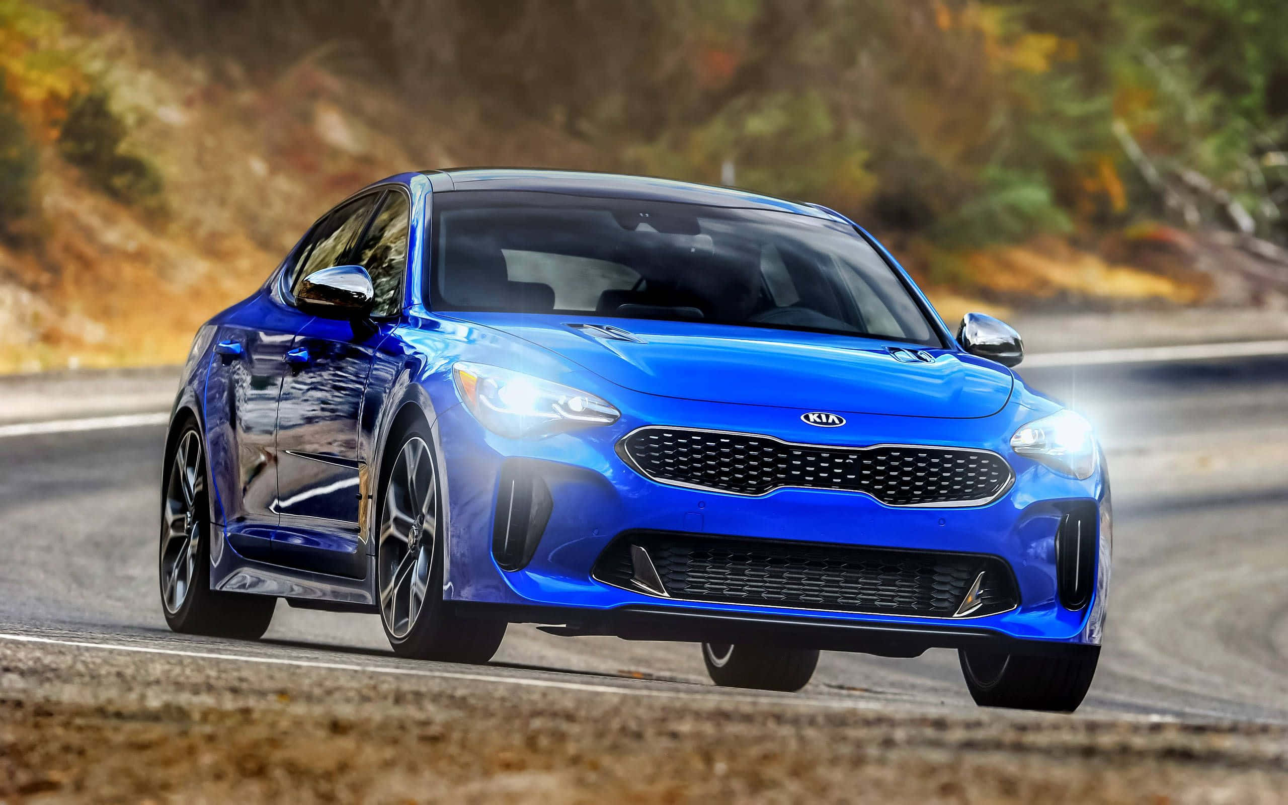 Kia Stinger 桌布 壁紙 — 一輛充滿活力的藍色Kia Stinger雙門跑車沿著蜿蜒的山路疾馳，背景是秋天的樹葉。