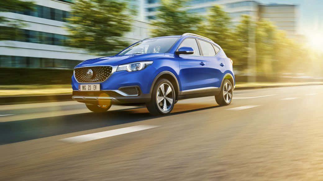  壁紙 — 一輛充滿活力的藍色MG ZS EV SUV在陽光明媚的城市道路上疾馳，背景有動態模糊和現代建築。
