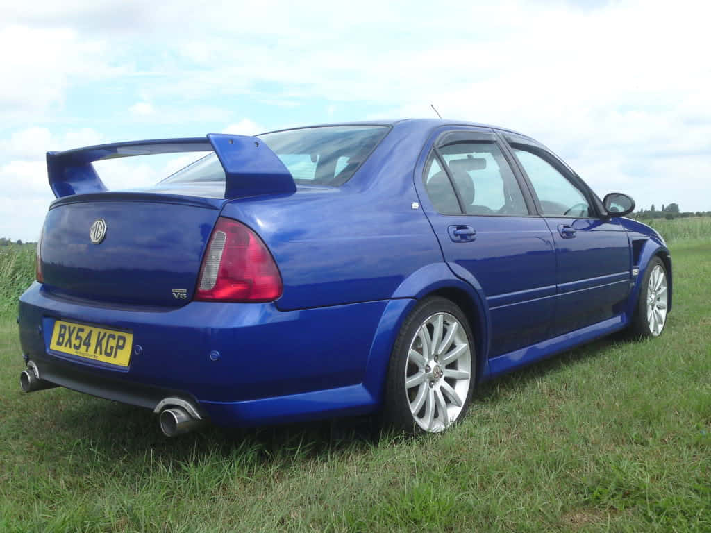  壁紙 — 一輛充滿活力的藍色MG ZT V6運動轎車，帶有大型後尾翼，停在部分多雲的天空下的草地上。