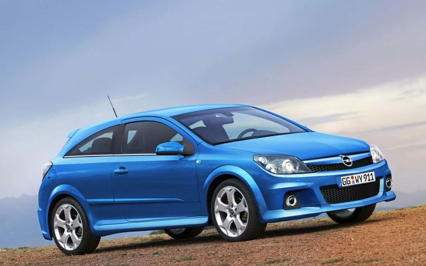Opel Astra 桌布 壁紙 — 一輛充滿活力的藍色歐寶雅特 GTC 跑車停泊在泥土山坡上，天空呈現柔和暮色。