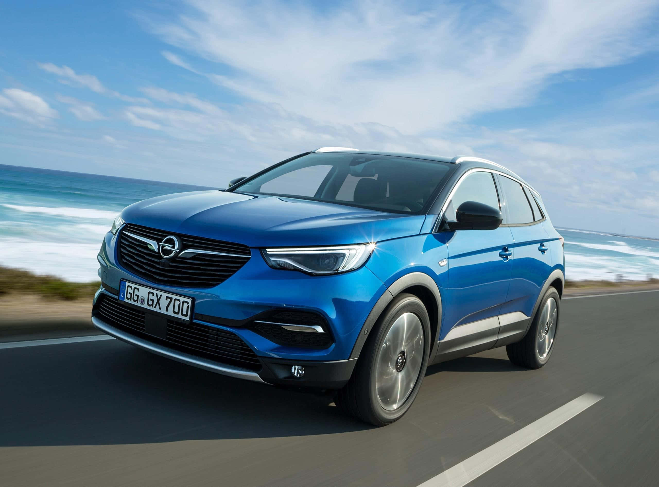 Opel Grandland X 桌布 壁紙 — 一輛充滿活力的藍色歐寶SUV沿著海岸公路行駛，海浪在明亮的藍天下拍打。