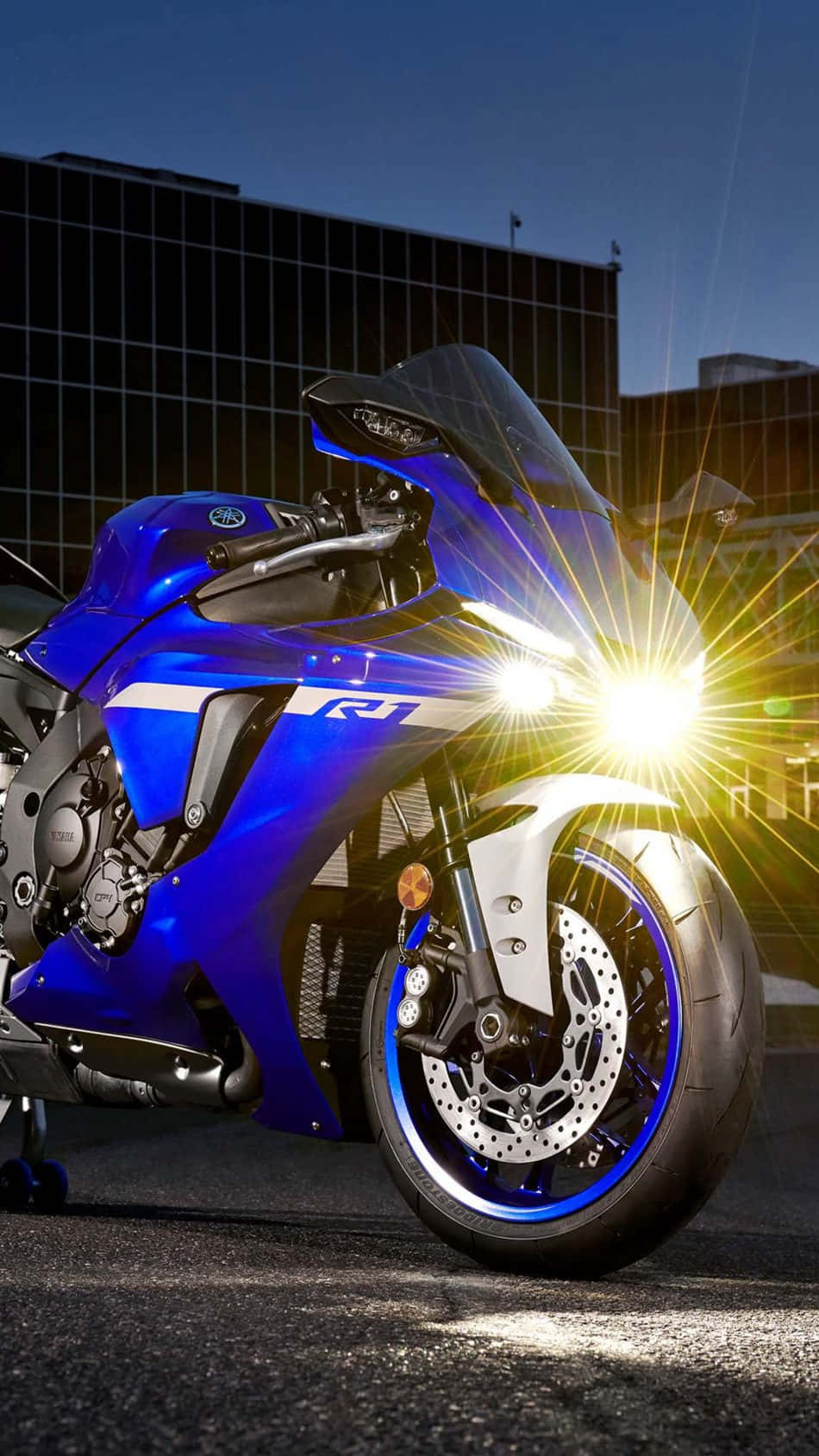  壁紙 — 一輛充滿活力的藍色運動電單車，帶有發光頭燈，在黃昏時停泊在現代建築前的瀝青路上。
