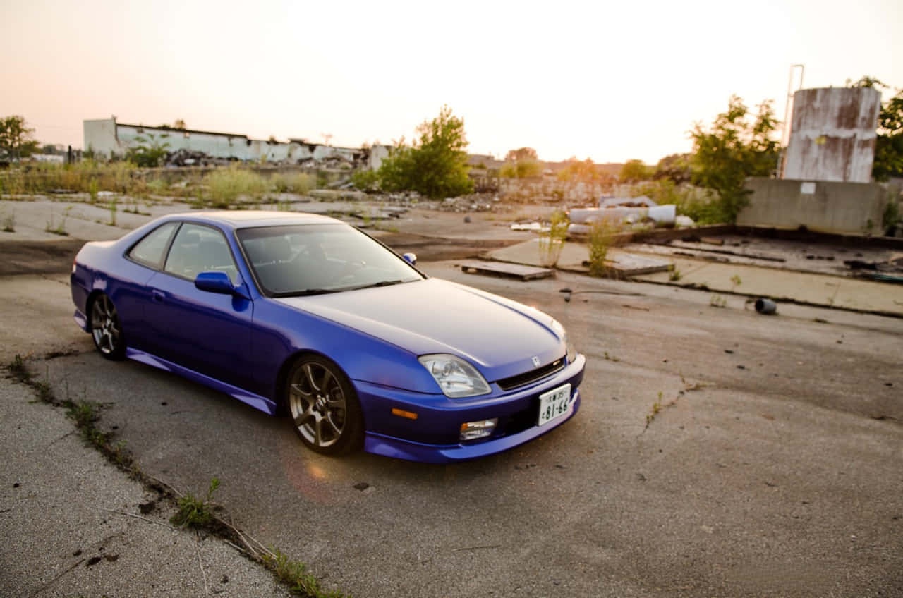 Honda Prelude 桌布 壁紙 — 一輛充滿活力的藍色跑車在黃金時段的光線下閃閃發光，獨自停在佈滿裂縫的混凝土上，周圍是雜草叢生的廢墟。