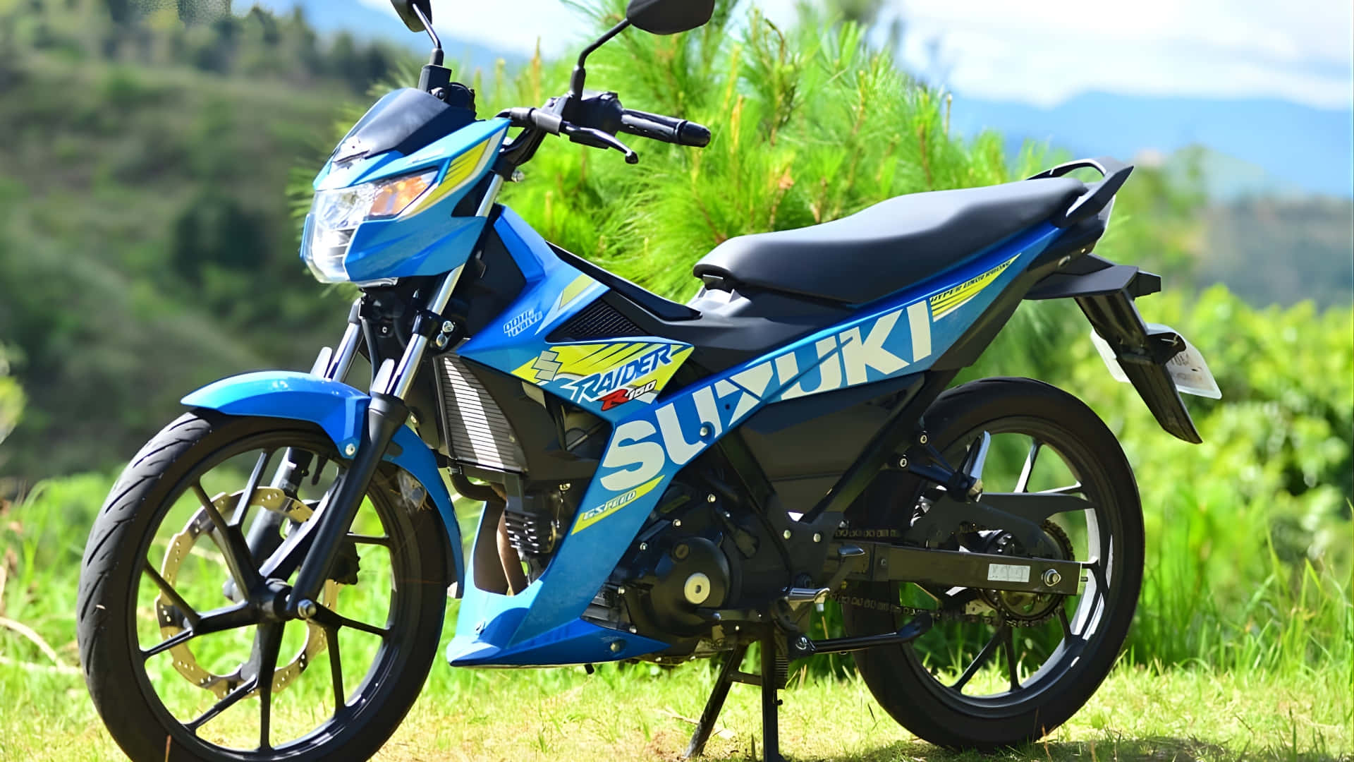  壁紙 — 一輛鮮藍色的Suzuki Raider 150電單車停在草地上，背景是翠綠的山丘和遠處的山脈，天空明亮。