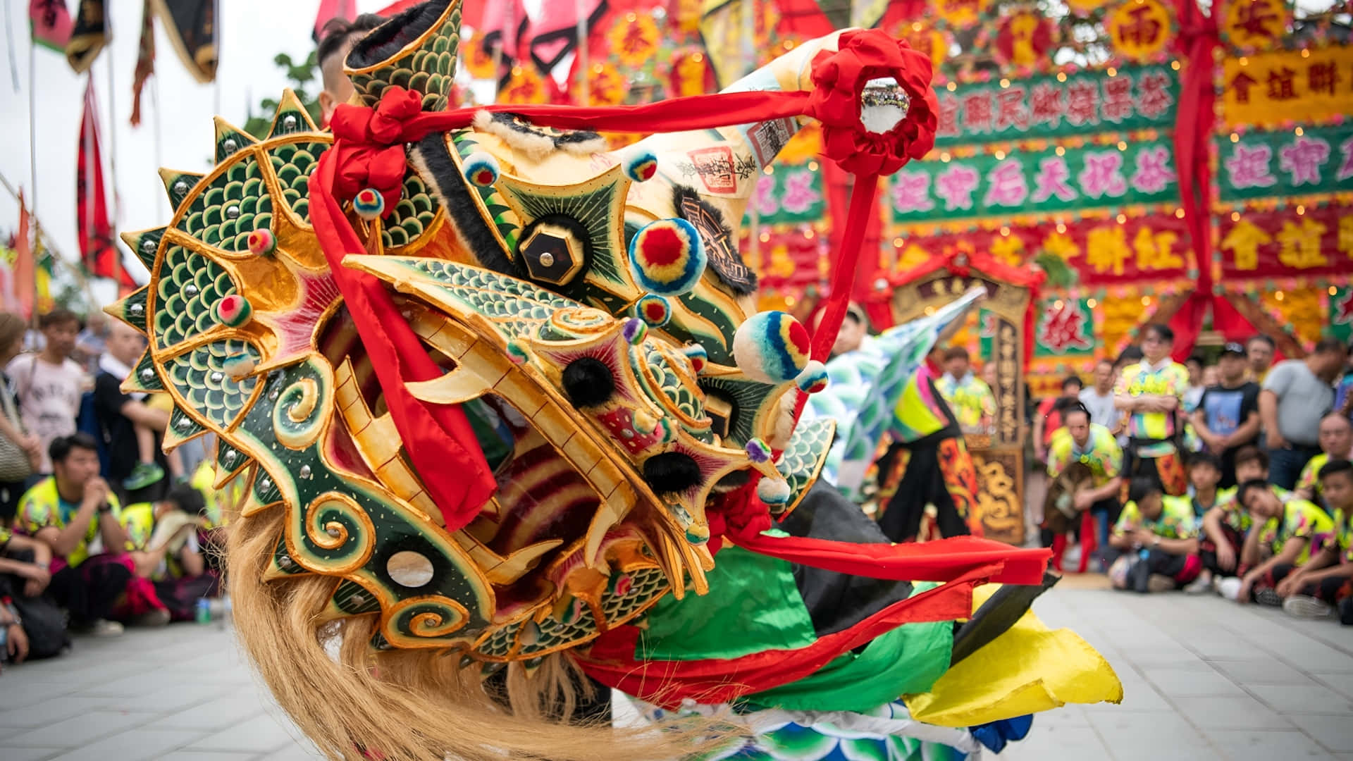  壁紙 — 一件充滿活力的舞龍服裝，帶有綠色、金色和紅色的細節，在擁擠的節日街道上充滿活力地移動。