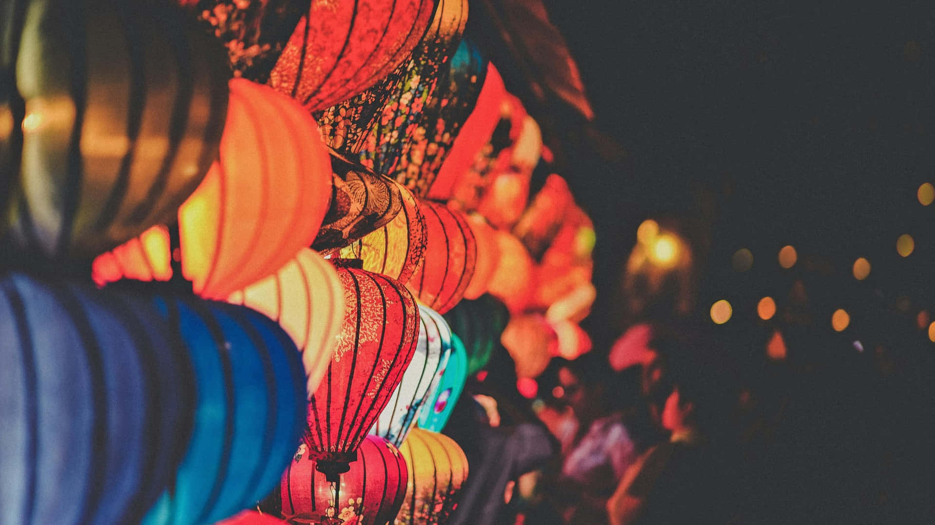  壁紙 — 紅色、藍色和金色的鮮豔發光燈籠在夜間密集懸掛，將溫暖的光線投射在下方模糊的人群上。