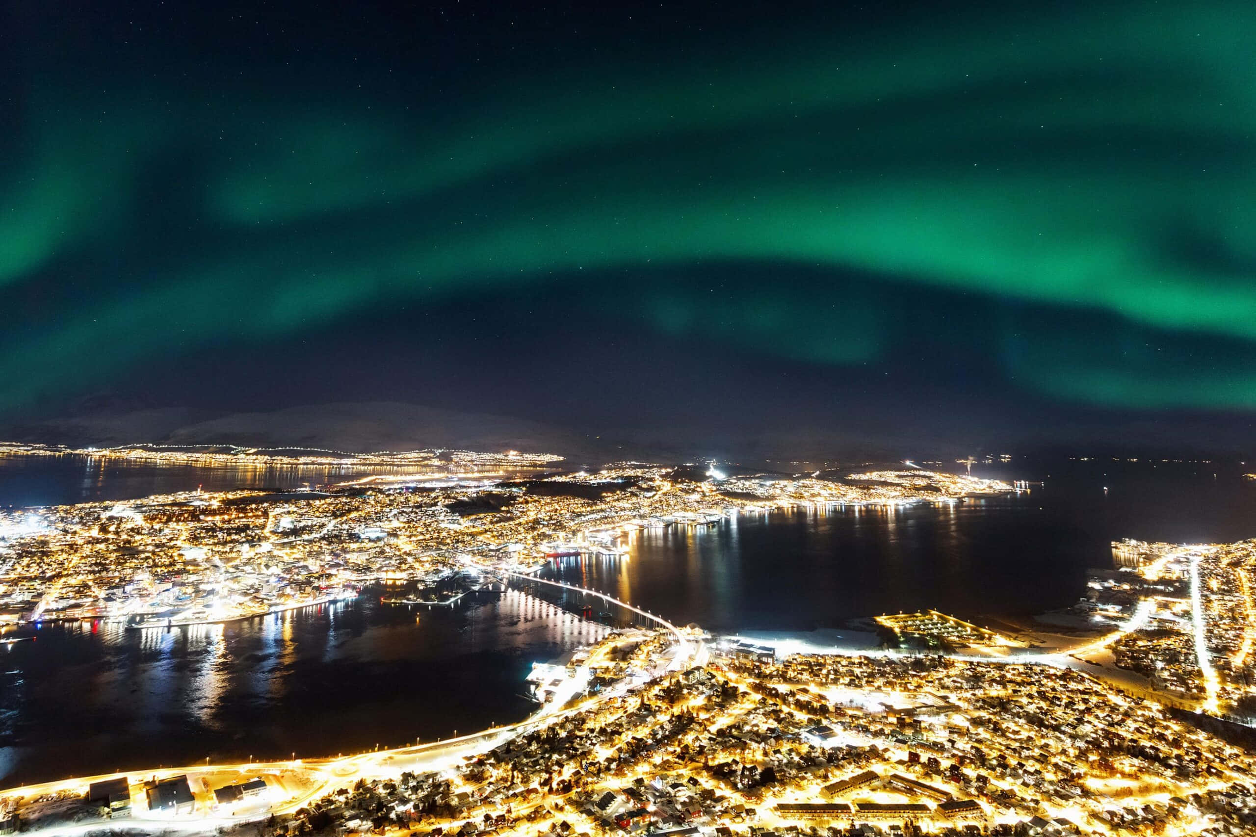 Tromsø 桌布 壁紙 — 充滿活力的綠色北極光在夜晚閃爍的沿海城市上空舞動，倒映在黑暗的水面上。