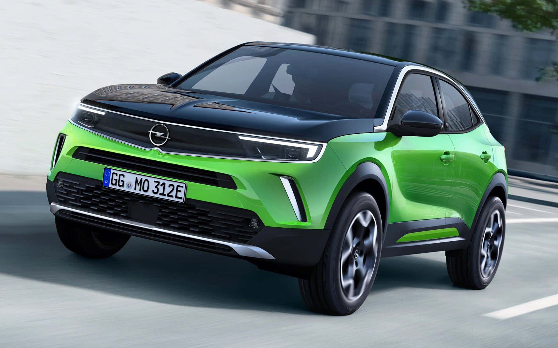 Opel Mokka 桌布 壁紙 — 一輛充滿活力的綠色Opel電動SUV在城巿街道上疾馳，背景有動態模糊和現代建築。