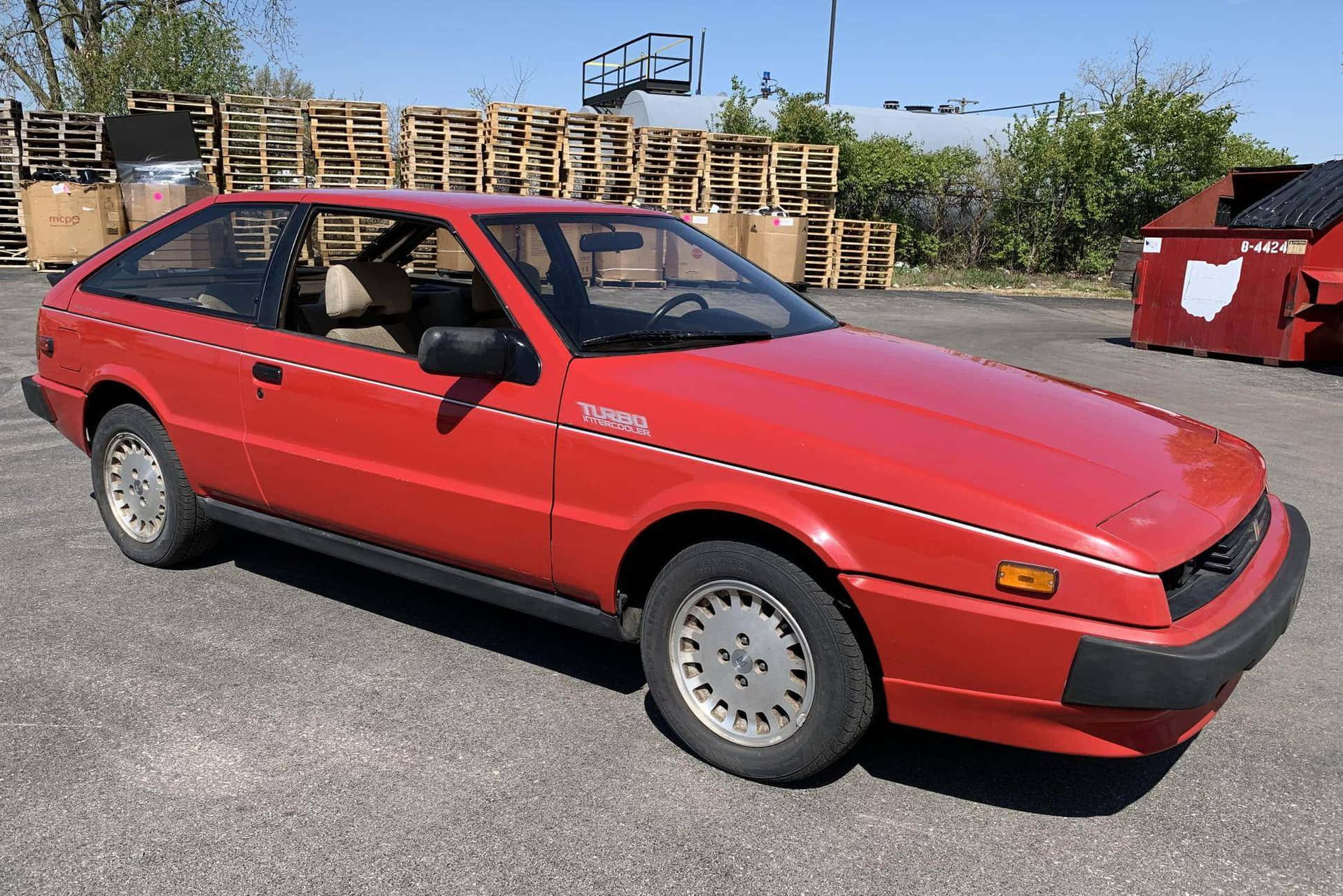  壁紙 — 一架鮮紅色嘅1980年代渦輪增壓掀背車停泊喺陽光普照嘅工業用地，背景有堆疊嘅木製卡板同一個紅色垃圾箱。