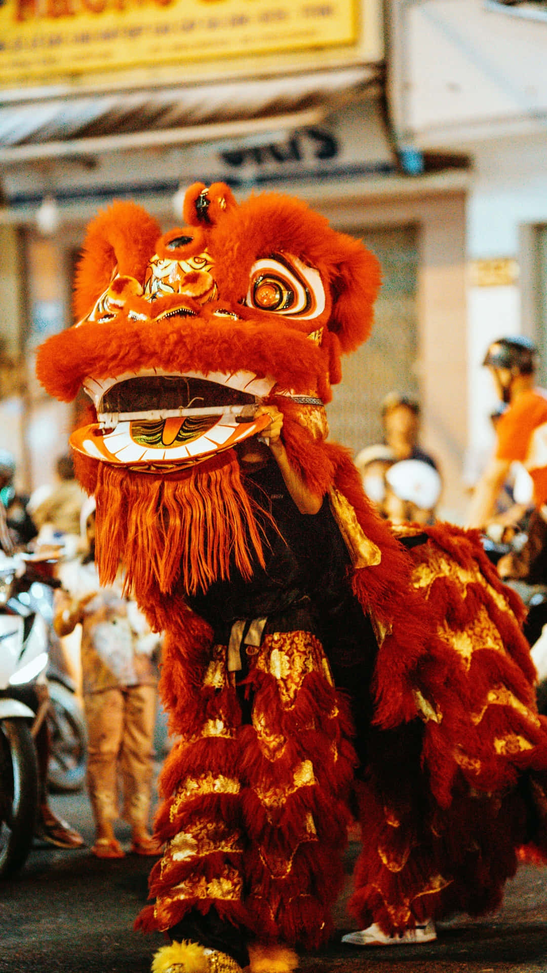  壁紙 — 一個充滿活力的紅金色舞獅服裝在繁忙的街道上充滿活力地表演，背景有模糊的觀眾和電單車。