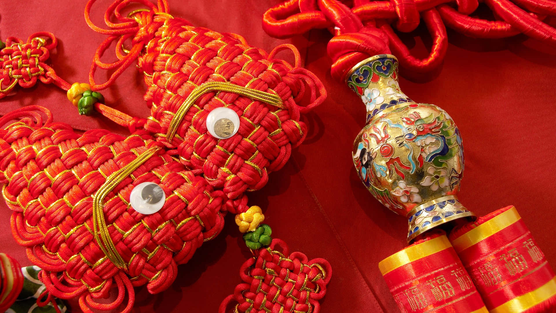  壁紙 — 鮮豔的紅色中國結魚飾和一個色彩繽紛的景泰藍花瓶放在豐富的紅色布料背景上。