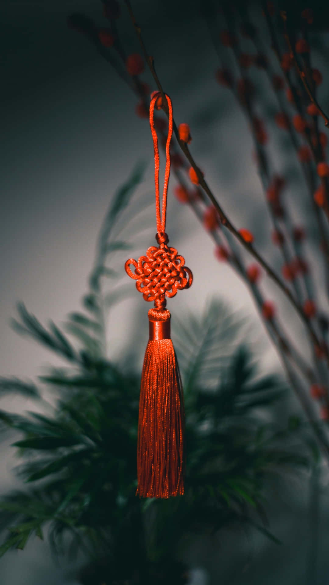  壁紙 — 一個鮮豔的紅色中國結飾品（帶流蘇）懸掛在模糊的綠色樹葉和掛滿漿果的樹枝前。