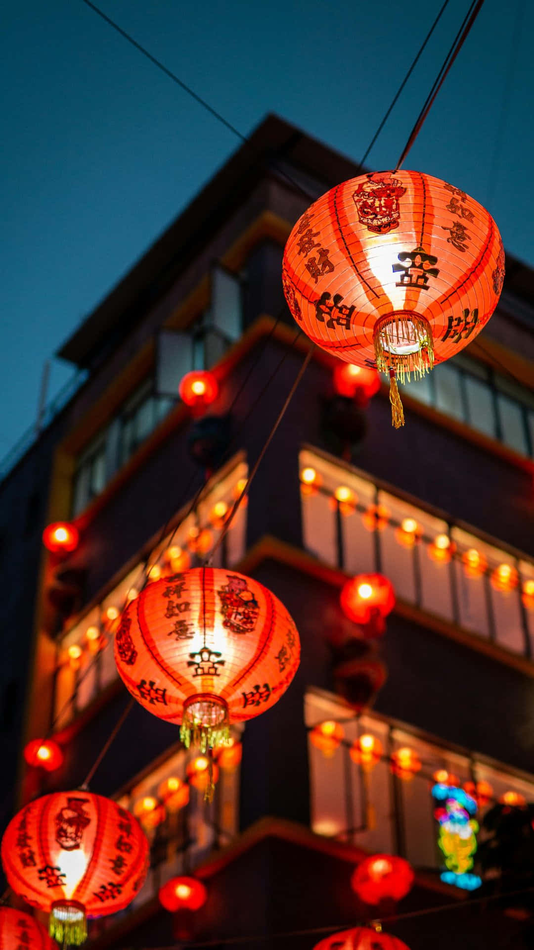  壁紙 — 鮮豔的紅色中國燈籠在深沉的暮色天空中溫暖地發光，上方是一座裝飾著串燈的建築物。
