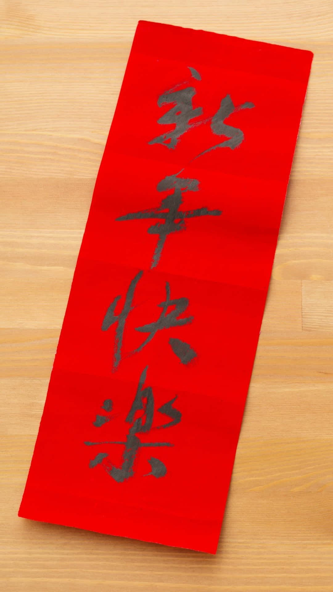  壁紙 — 一幅鮮紅色的中國新年捲軸，上面有黑色書法寫著「新年快樂」，放在淺色木質表面上。