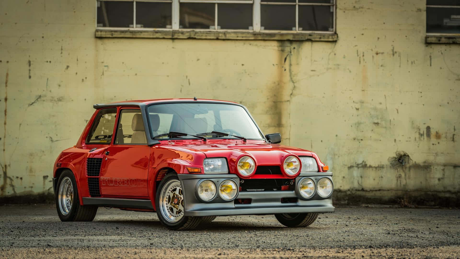 Renault 5 Turbo 桌布 壁紙 — 一輛鮮紅色的經典拉力賽車，帶有六個圓形頭燈，停在一座風化的工業建築前。