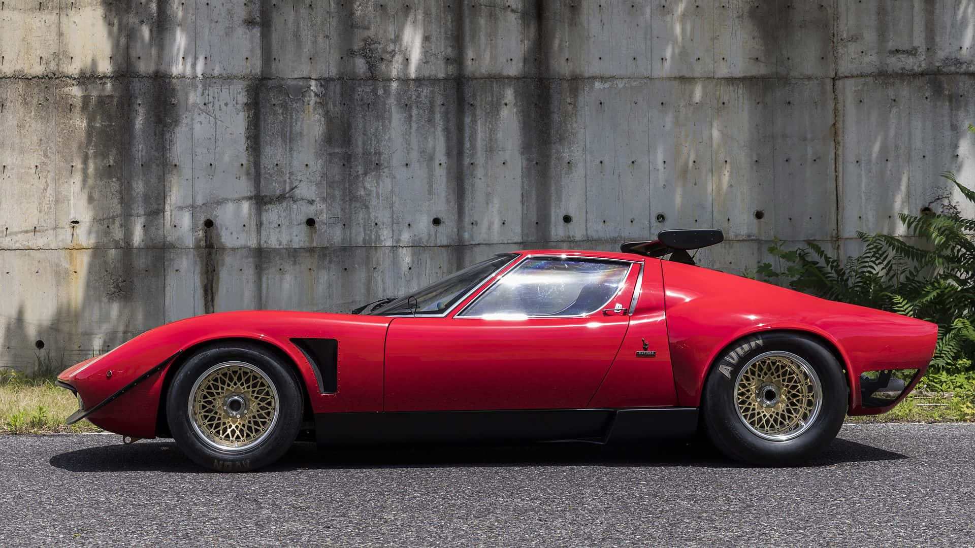 Lamborghini Miura 桌布 壁紙 — 一輛鮮紅色經典跑車配金色鋼絲輪圈，停泊在風化的混凝土牆旁。