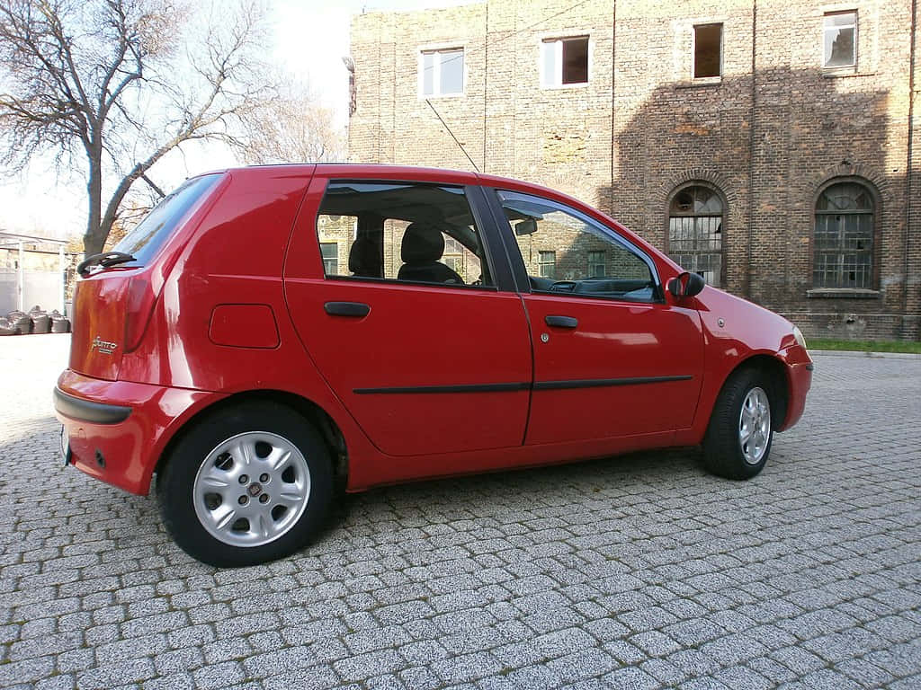 Fiat Punto 桌布 壁紙 — 一輛鮮紅色嘅小型車停泊喺鵝卵石庭院，旁邊係一座受風化嘅磚砌建築，喺晴朗嘅日光下。