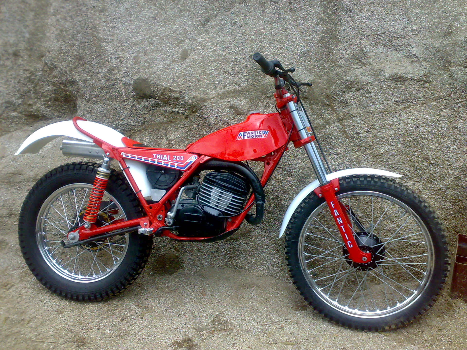  壁紙 — 一架鮮紅色嘅Fantich Trial 200越野電單車喺碎石地上靠住粗糙嘅石牆企定。