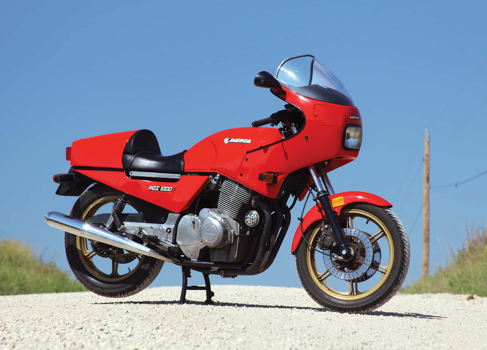  壁紙 — 一架鮮紅色嘅Honda RGS 1000電單車喺晴朗藍天下嘅碎石路上蓄勢待發，背景有綠色草地。