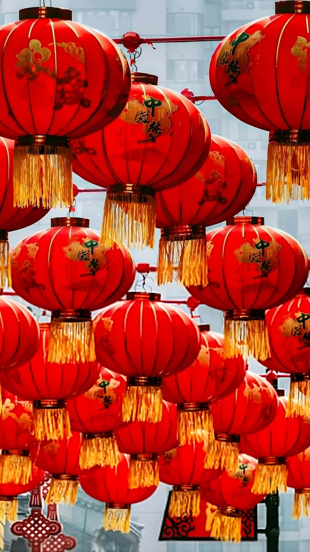  壁紙 — 鮮豔的紅色燈籠，帶有金色流蘇和漢字，密集地懸掛在模糊的城市背景前。