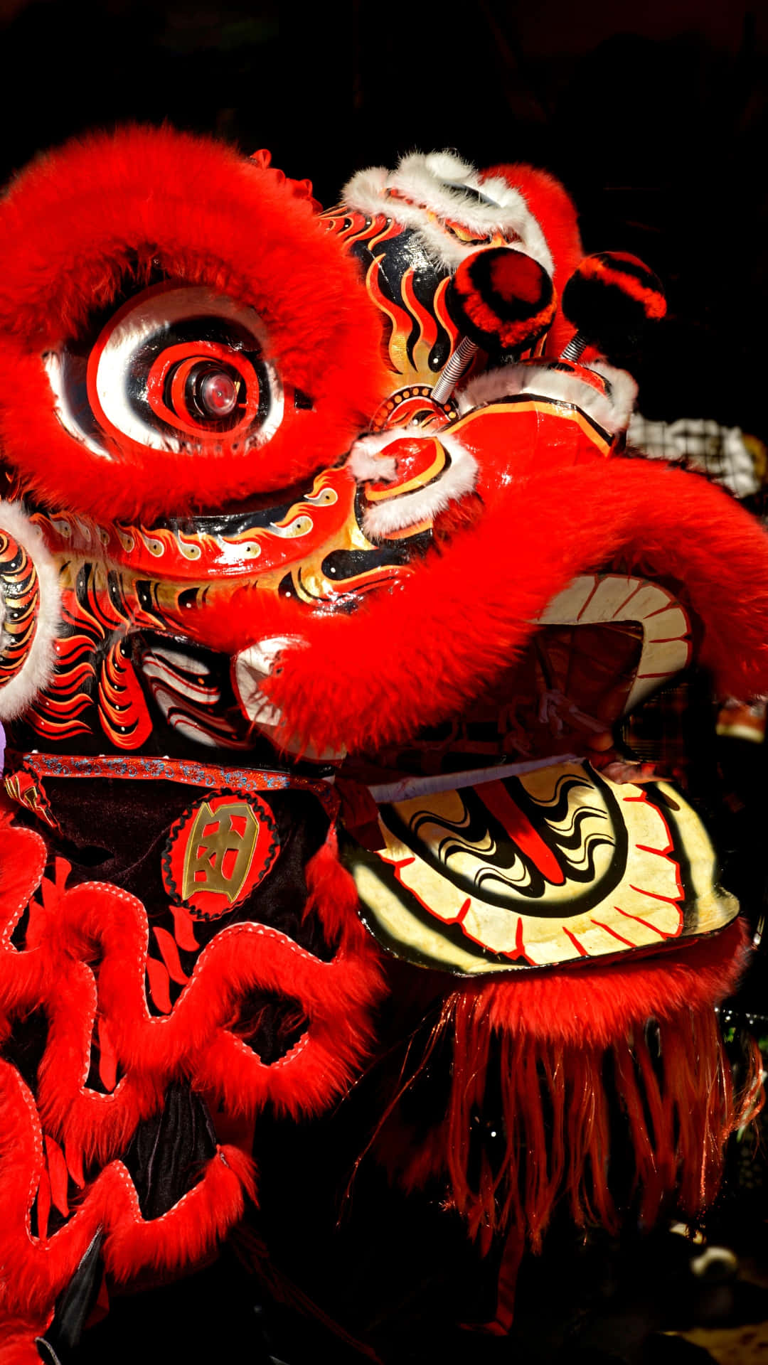  壁紙 — 一件鮮紅色嘅舞獅服裝，帶有複雜嘅黑色同金色圖案，喺戲劇性嘅舞台燈光下閃耀。