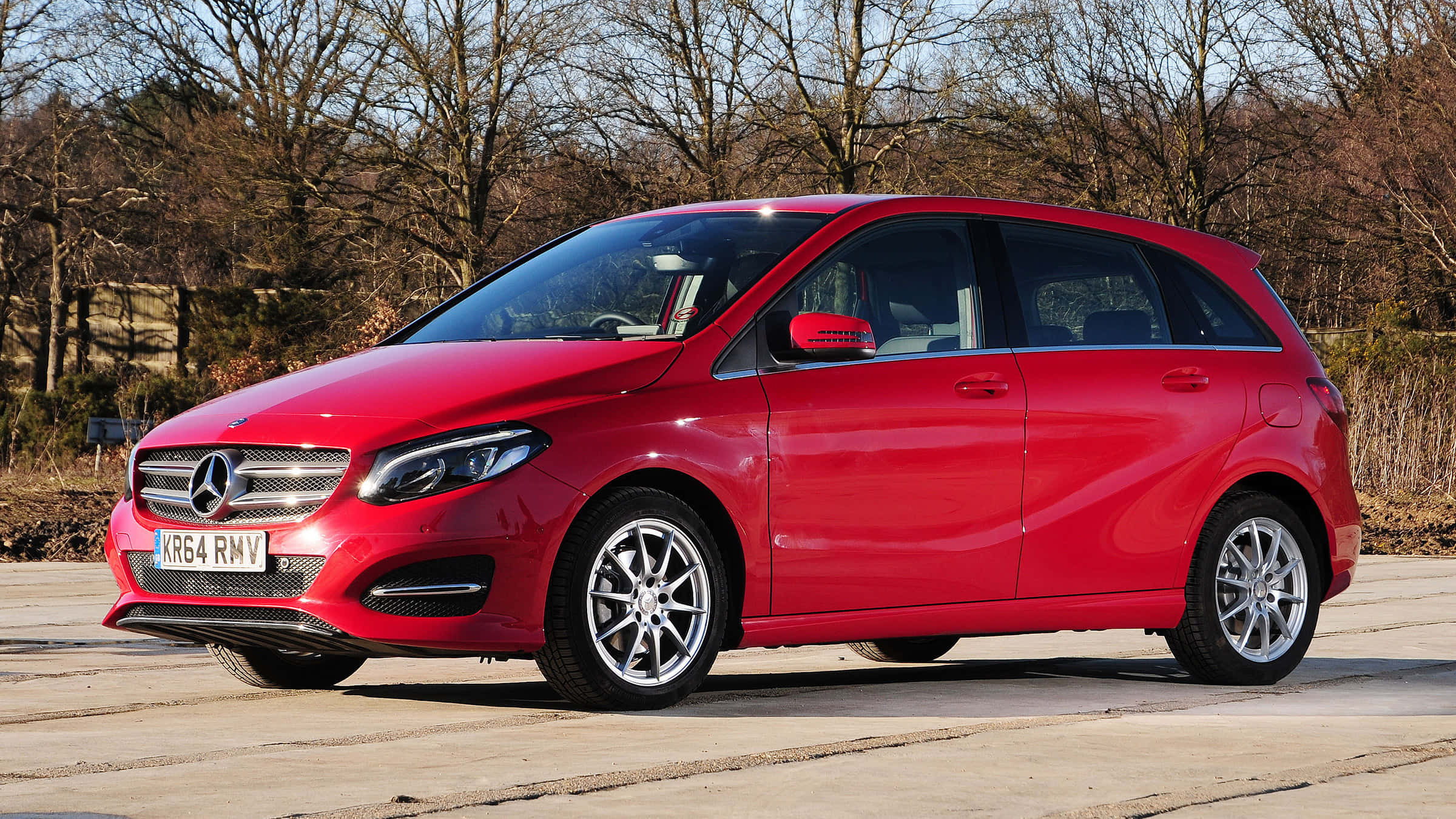 Mercedes Benz B-class 桌布 壁紙 — 一架鮮紅色嘅Mercedes-Benz B-Class掀背車停泊喺鋪砌嘅停車場，背景有光禿禿嘅樹木，喺晴朗嘅日光下。