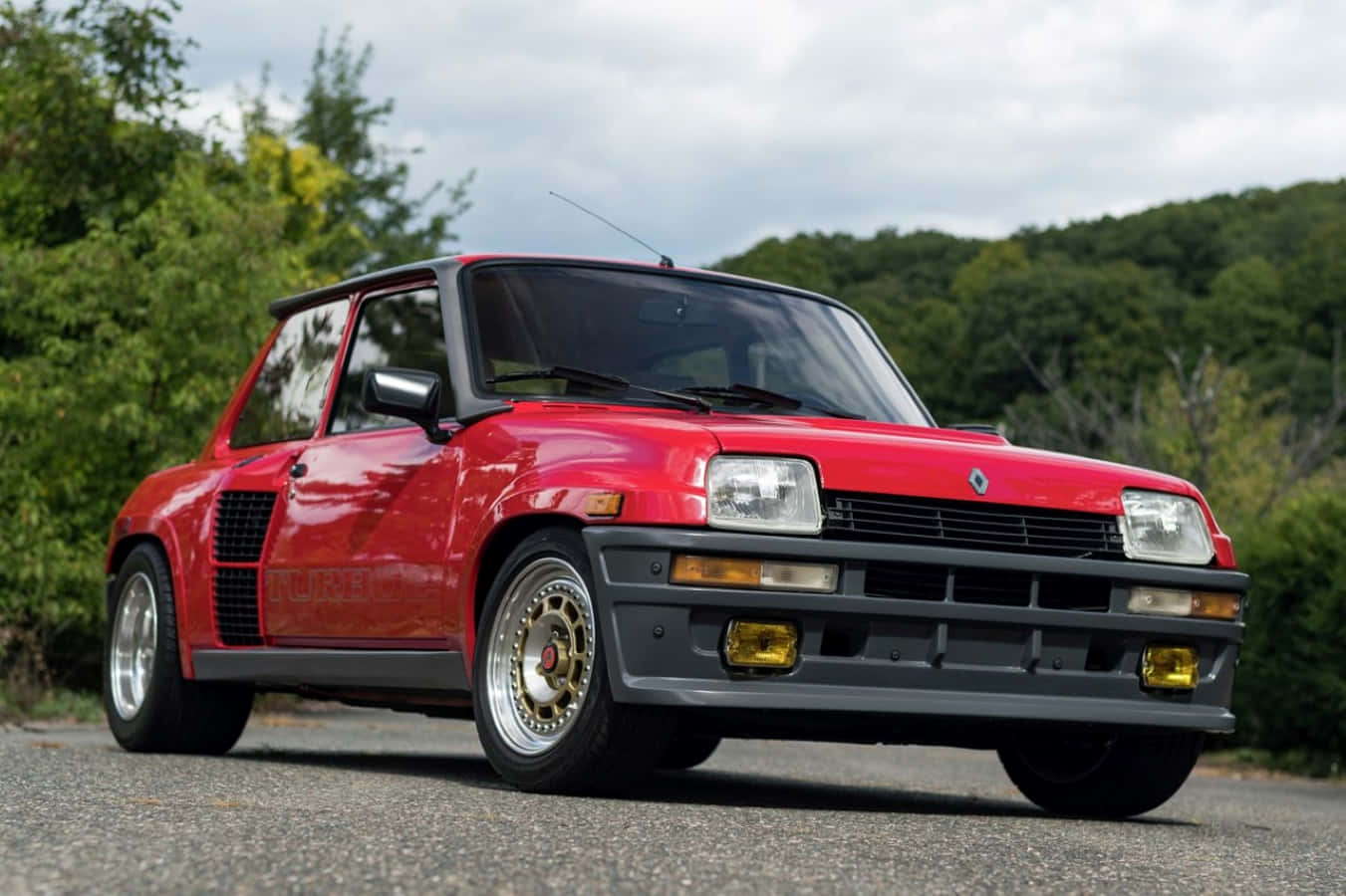 Renault 5 Turbo 桌布 壁紙 — 一架鮮紅色的雷諾5 Turbo 2跑車停泊在碎石路上，背景是茂密的綠樹和山丘，天空多雲。