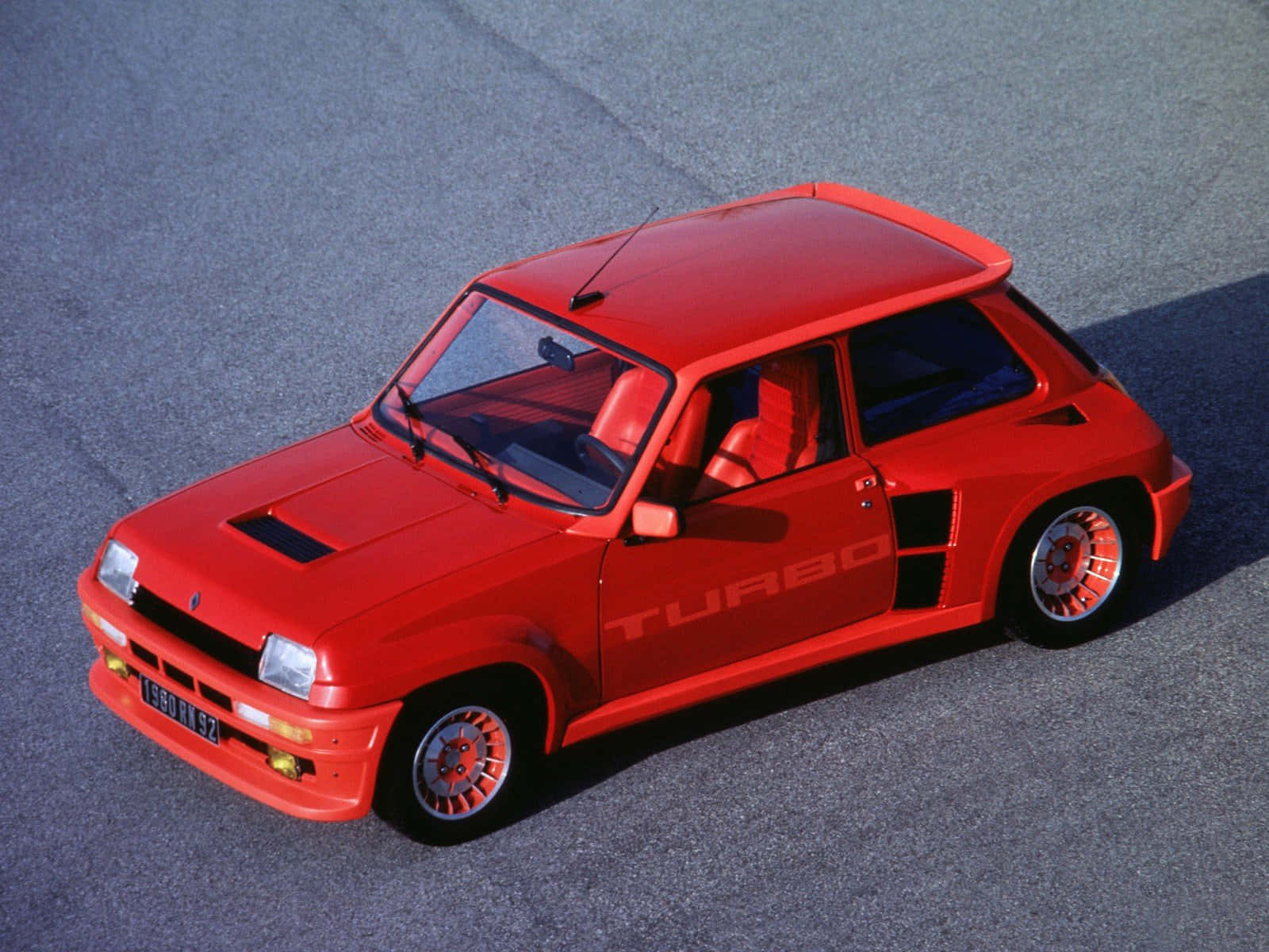 Renault 5 Turbo 桌布 壁紙 — 一輛鮮紅色的雷諾5 Turbo跑車在明亮的日光下停泊在灰色瀝青路面上。
