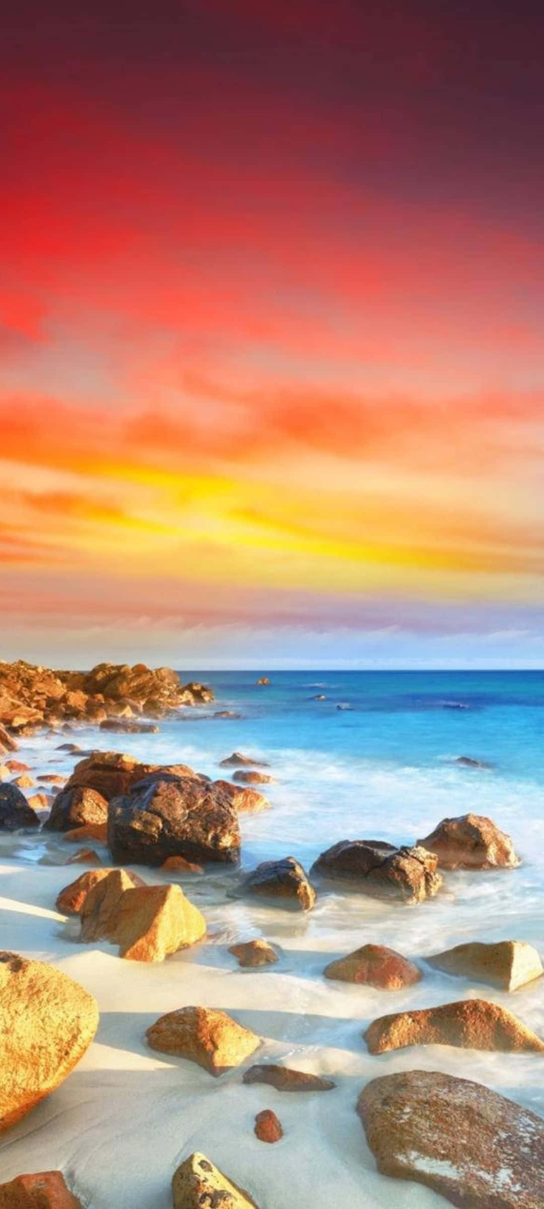 Pixel 7 桌布 壁紙 — 鮮豔的夕陽將天空染成火紅、橙黃色，照耀著岩石海灘，綠松石色的波浪拍打在金色沙灘上。