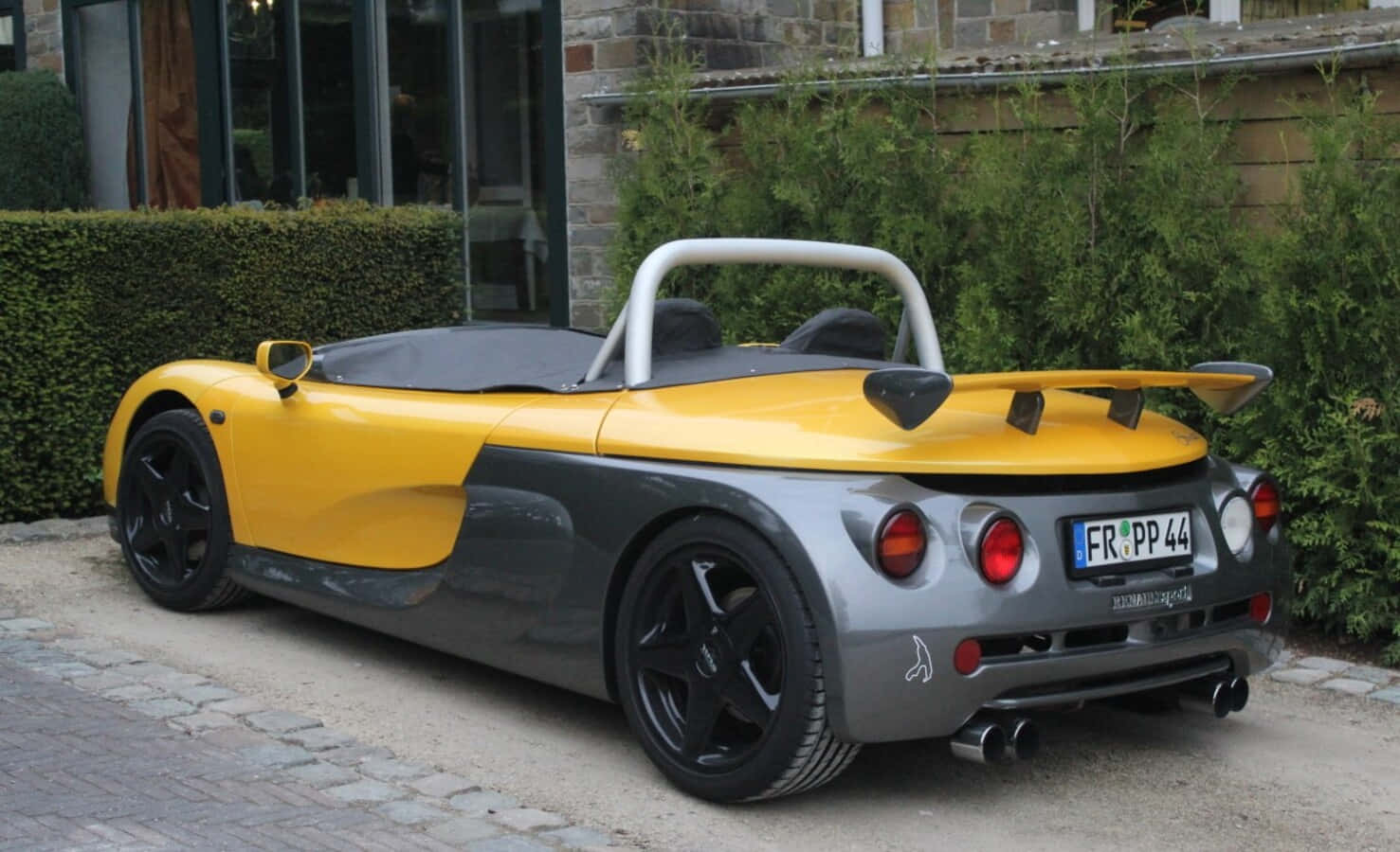 Renault Sport Spider 桌布 壁紙 — 一輛鮮黃色和灰色的跑車，帶有白色防滾架，停在鵝卵石車道上，旁邊是綠色樹籬。